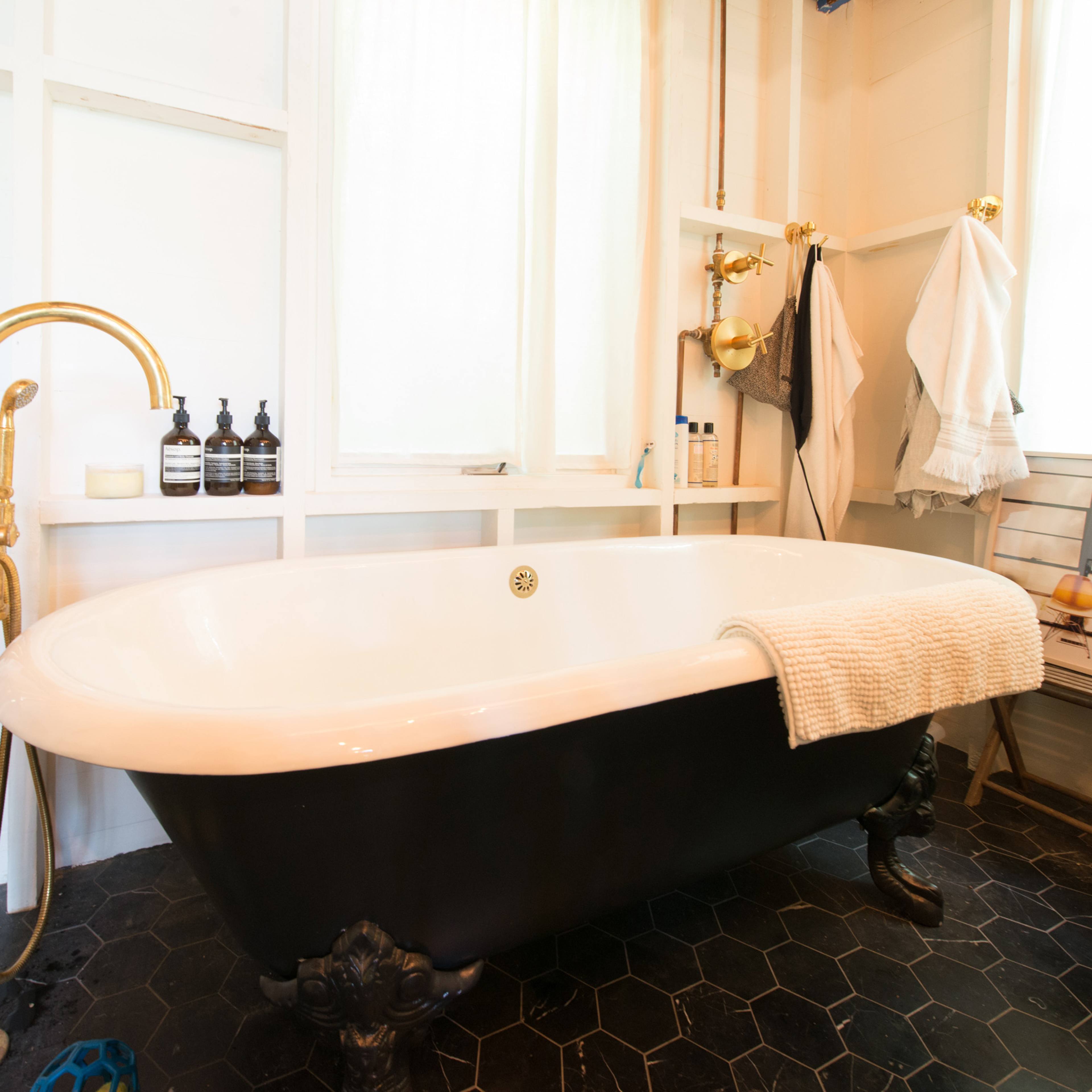 A black-and-white freestanding bathtub with a gold faucet in a well-lit bathroom featuring white walls and hexagonal tile flooring.