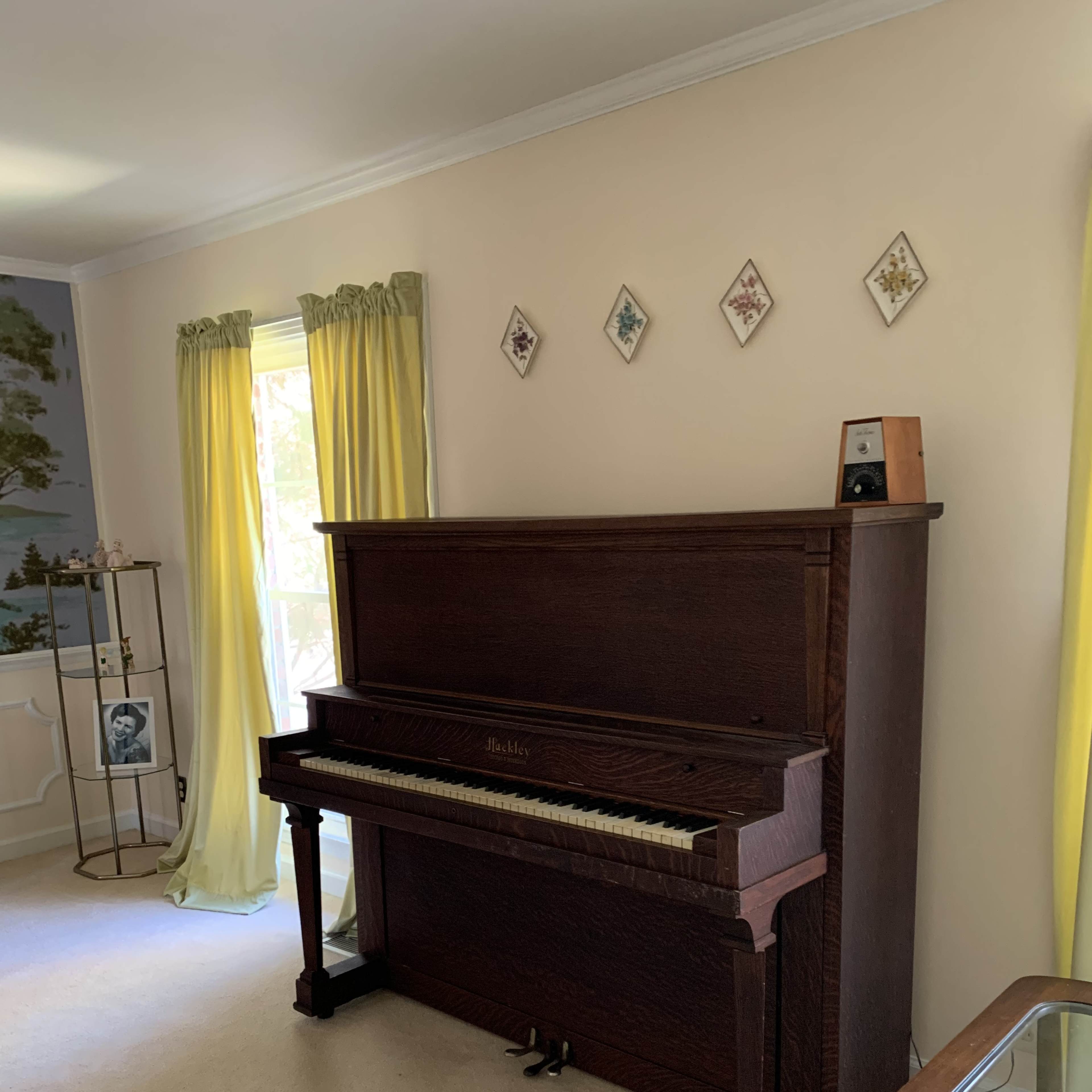 A wooden piano is positioned against a wall in a room with yellow curtains and decorative wall art above it.