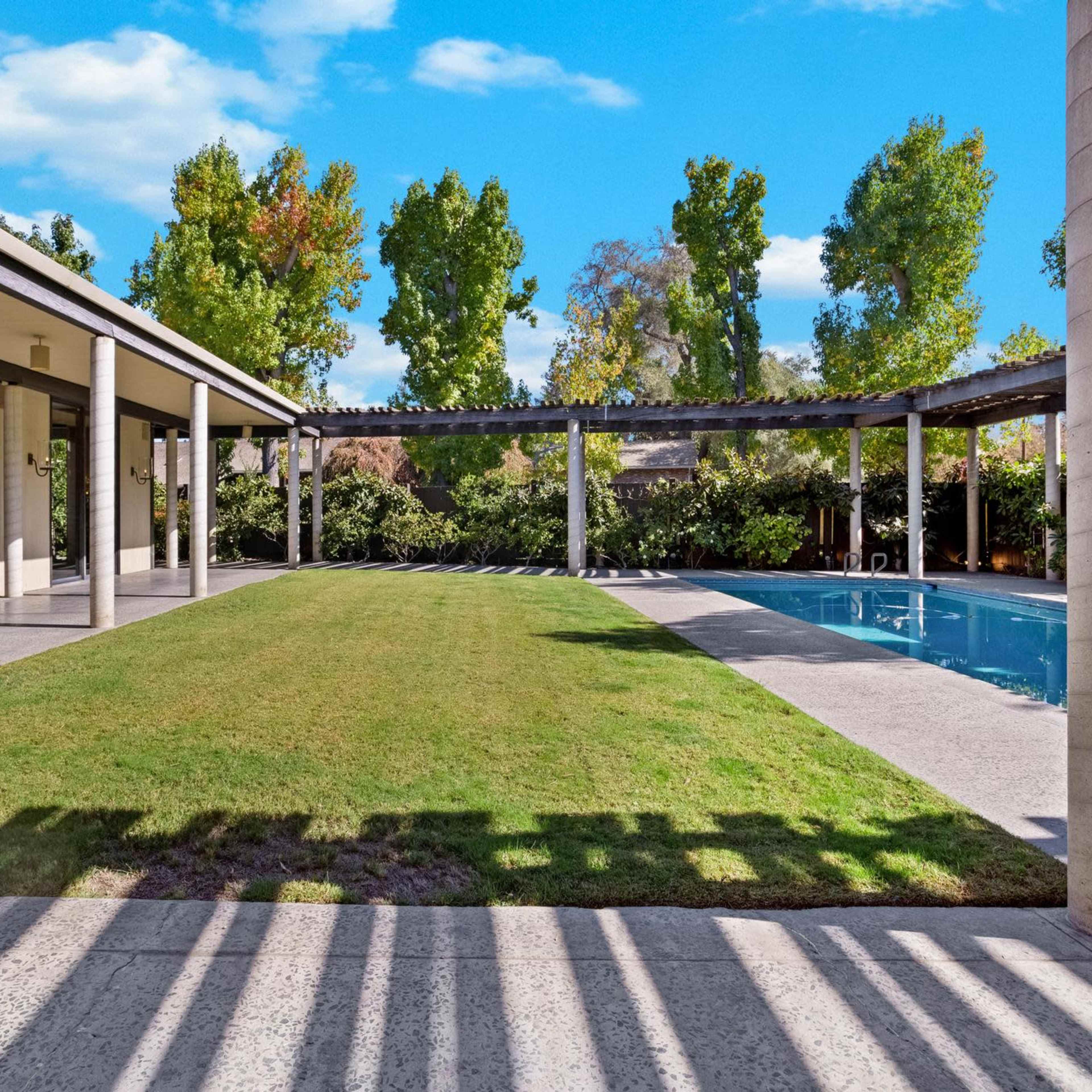 A landscaped backyard with a pool, green grass, and trees, framed by a modern house with a covered patio.