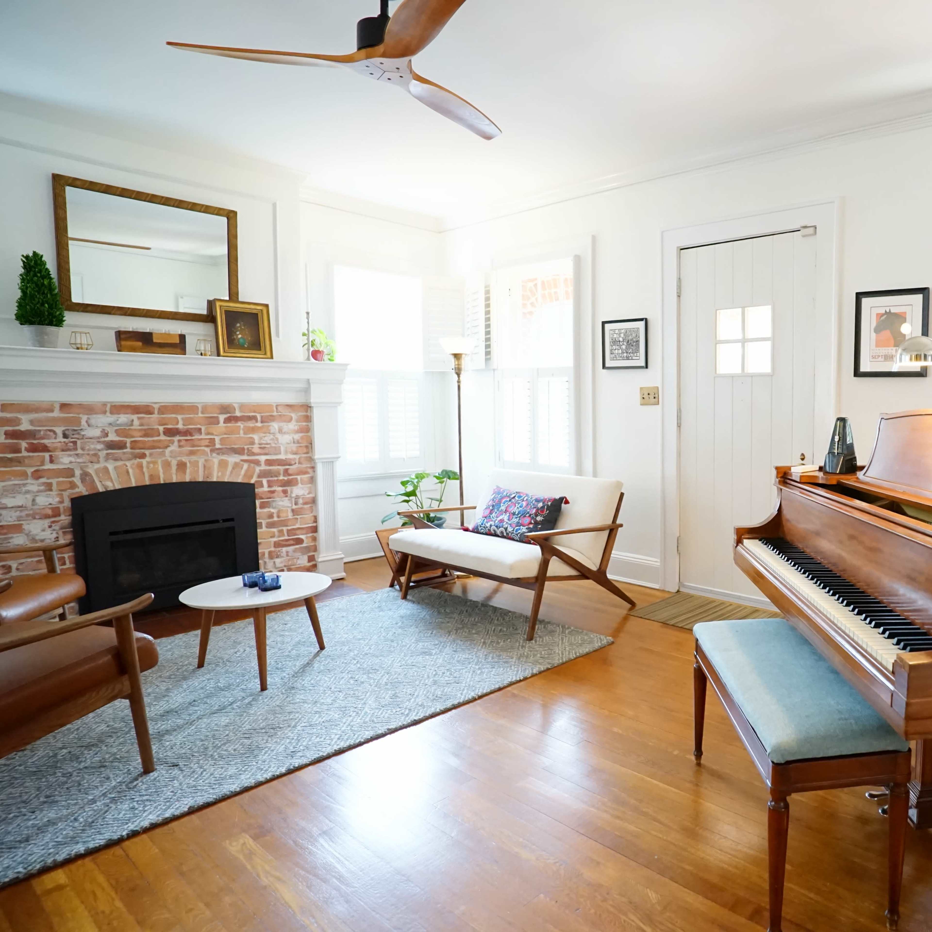 A cozy living room featuring a grand piano, a fireplace with exposed brick, a light-colored sofa, and a coffee table on a textured rug.