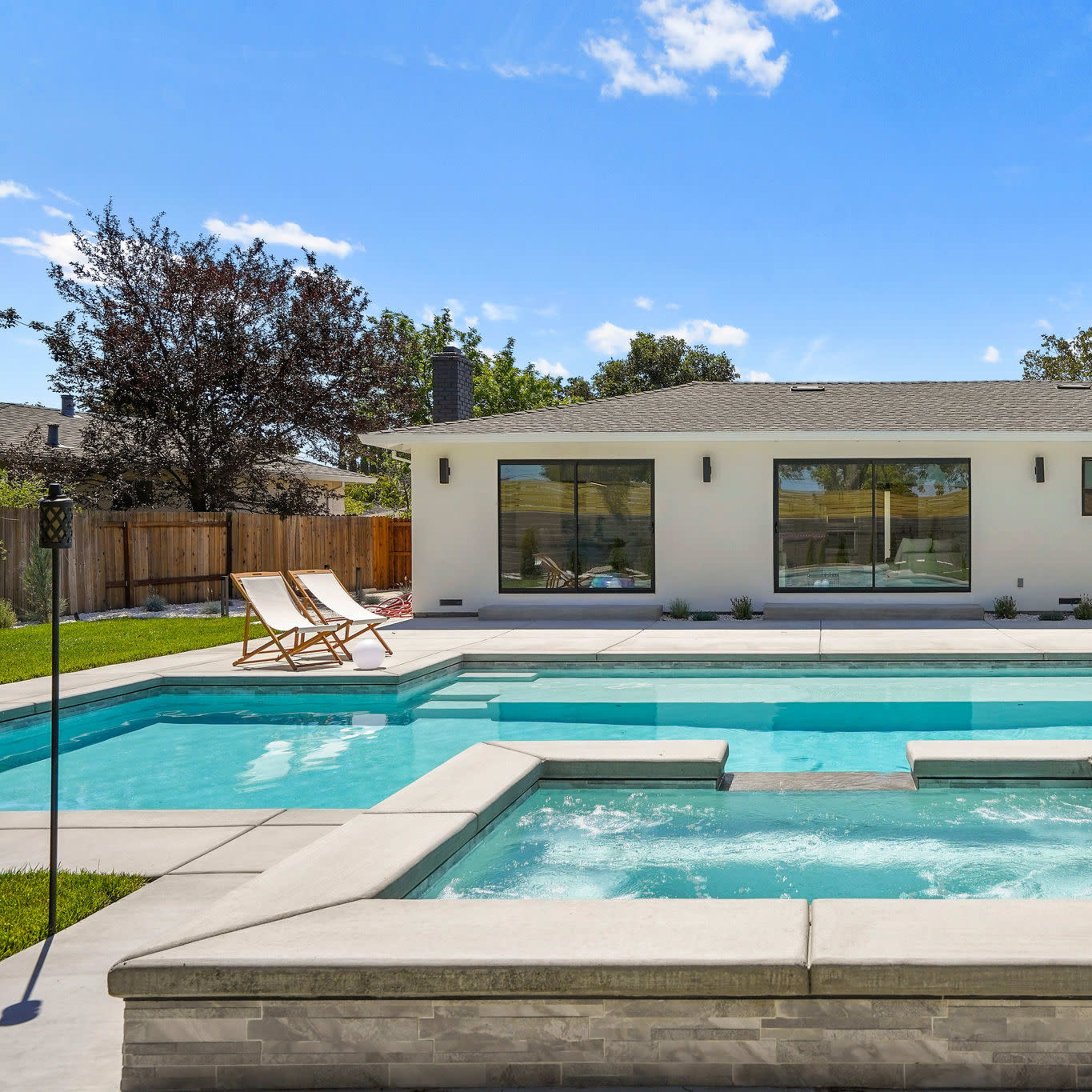 A modern home with large windows and a swimming pool featuring a spa area in the foreground surrounded by a landscaped yard.