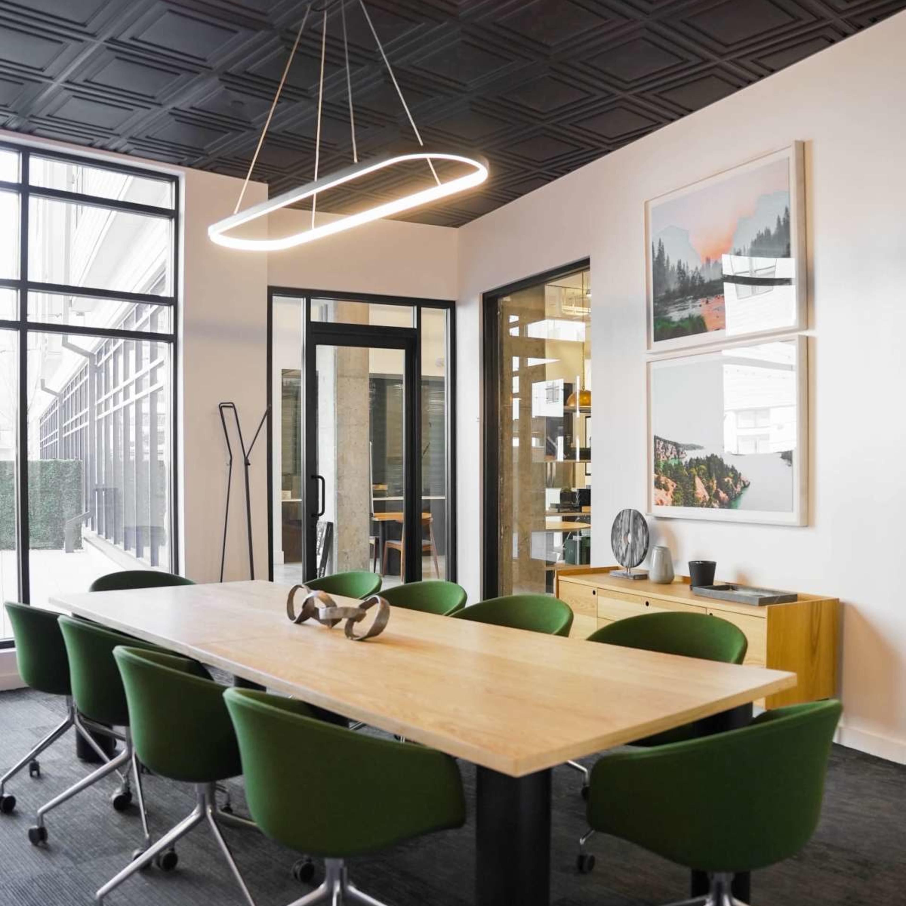 A modern conference room features a large wooden table surrounded by green rolling chairs, along with a wall displaying framed art.