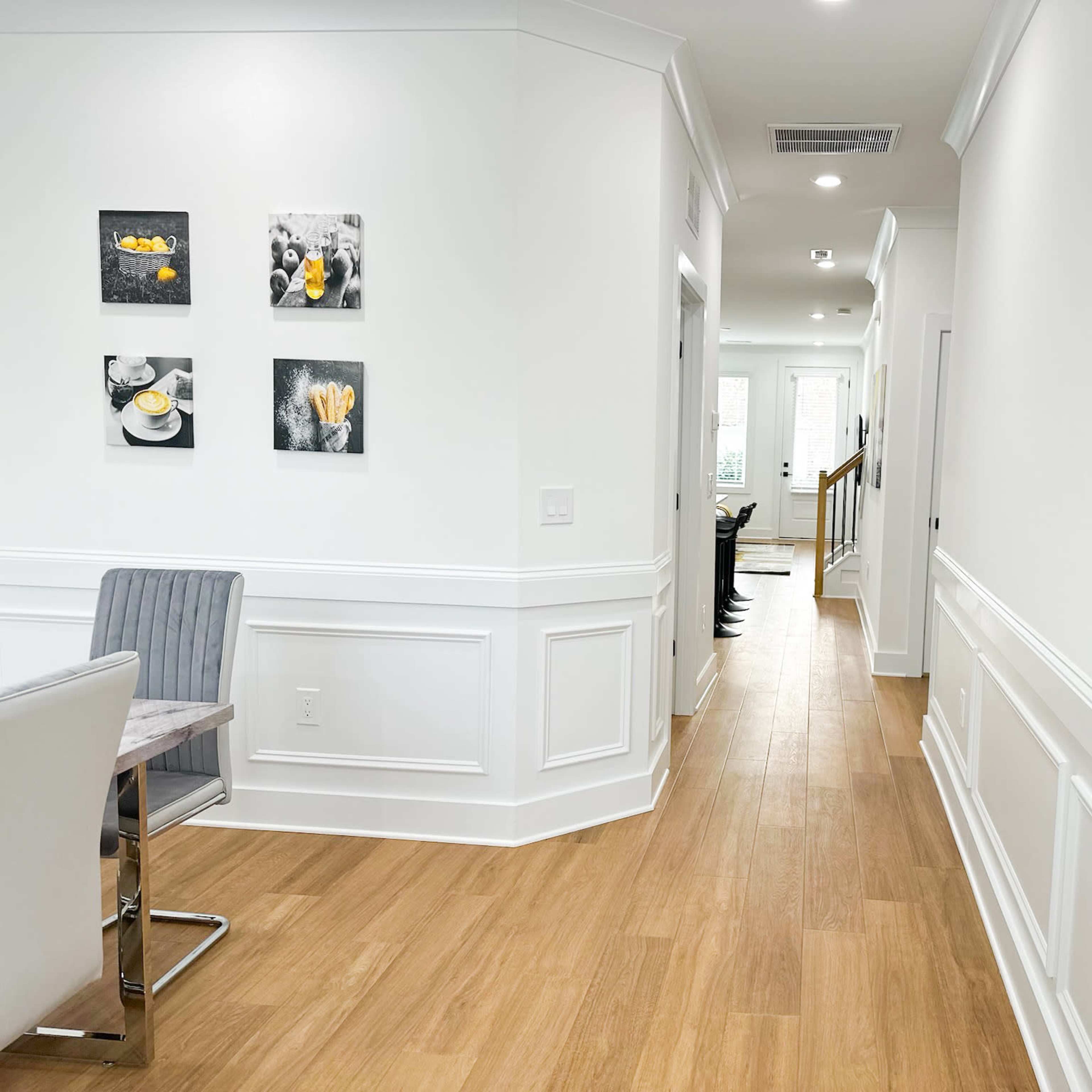 A bright corridor features white walls, wood flooring, and framed art on one side, leading to a staircase and open space at the end.