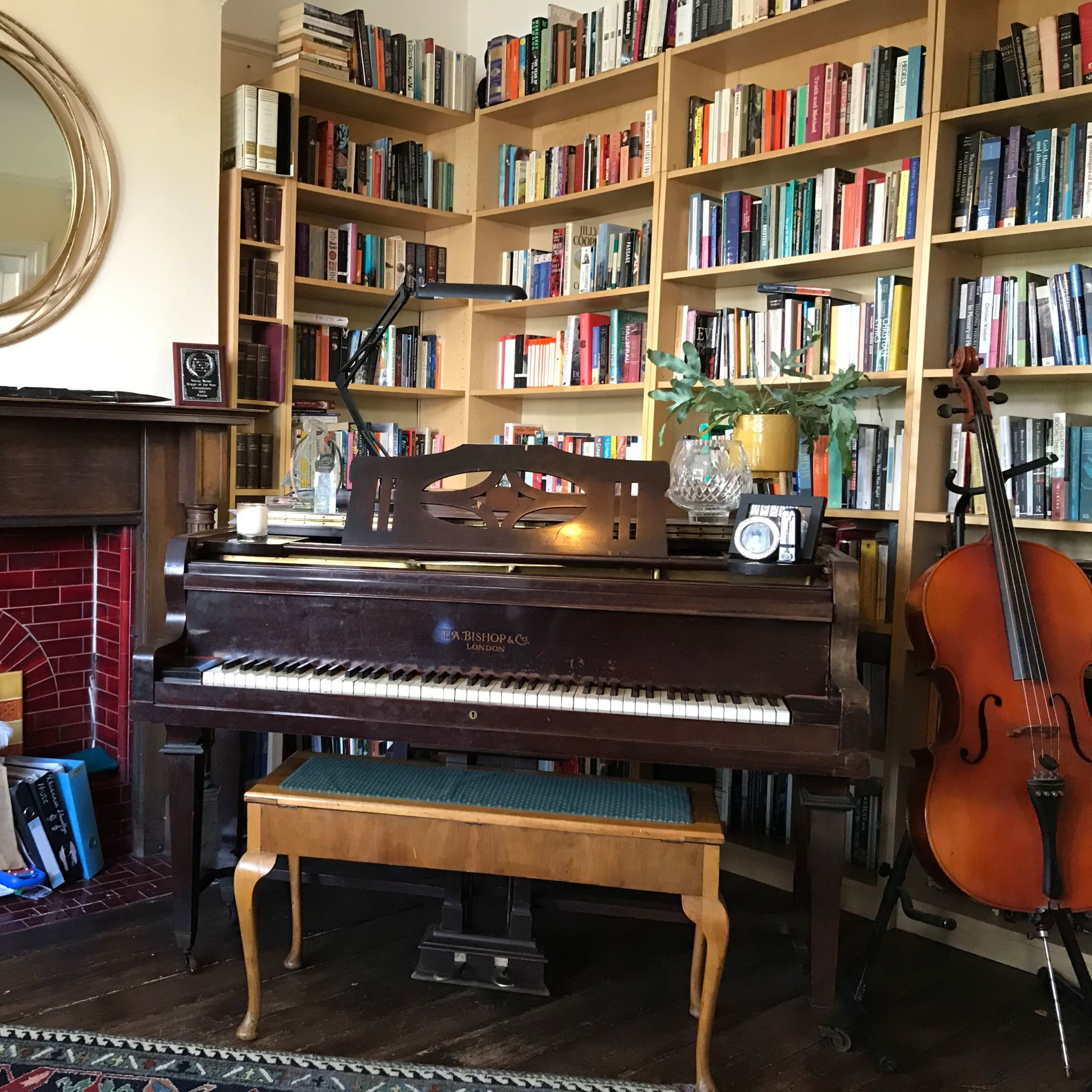A wooden piano is positioned next to a filled bookshelf in a cozy room, with a stack of books leaning against the fireplace and a cello standing nearby.