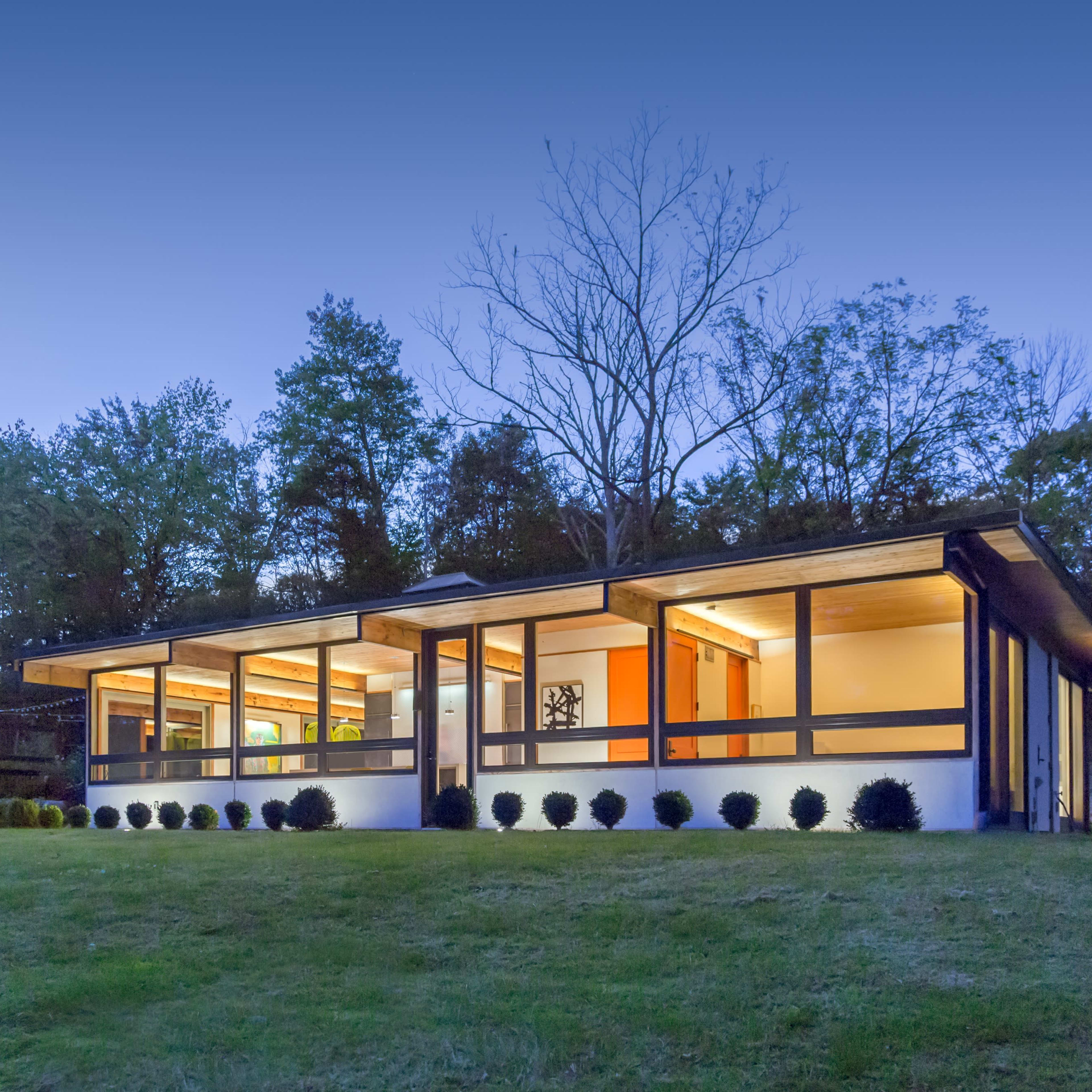 A modern house with large windows is set against a backdrop of trees in the evening light.