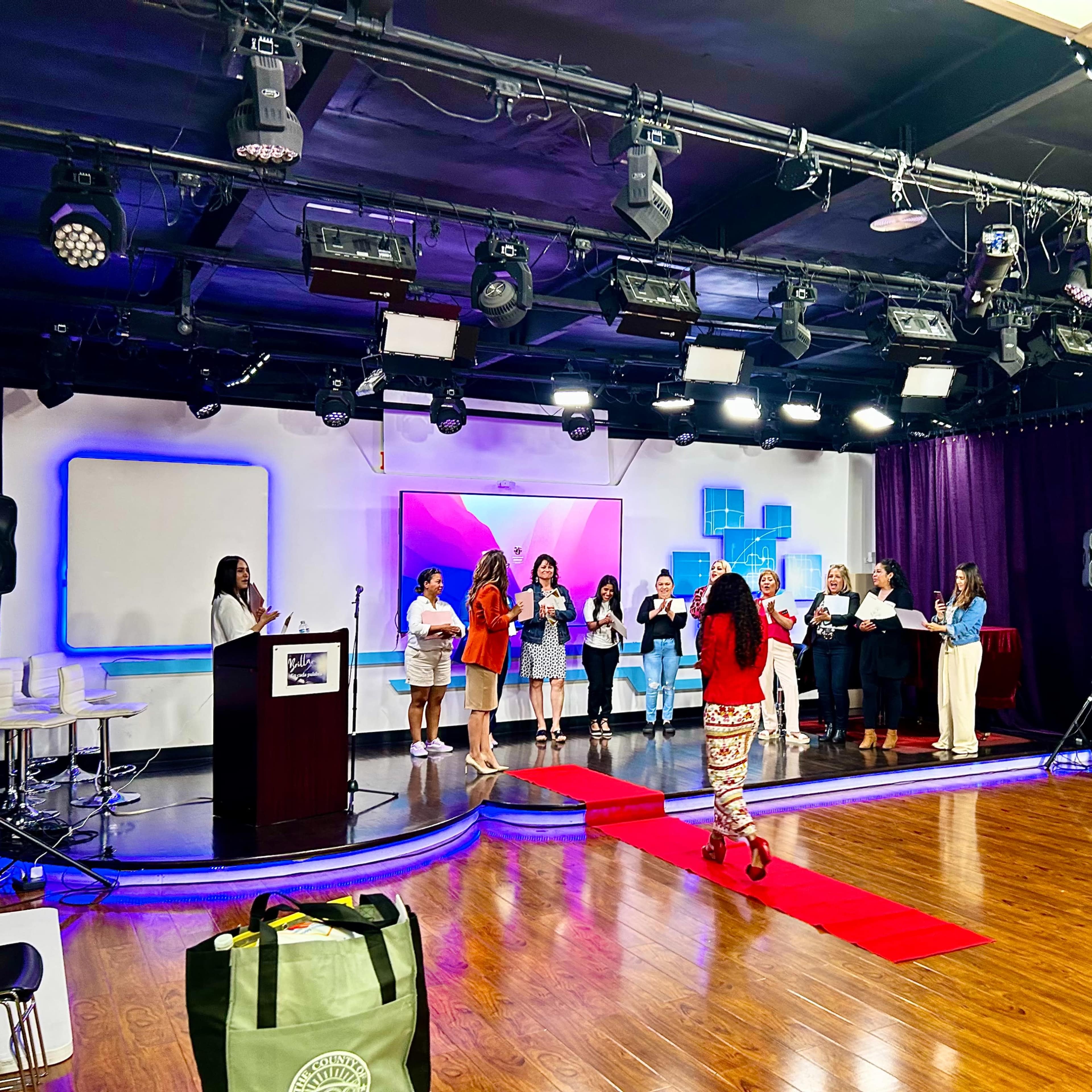 A group of people stands on a stage in a brightly lit room, with a red carpet leading to the front amidst a set of audio-visual equipment.