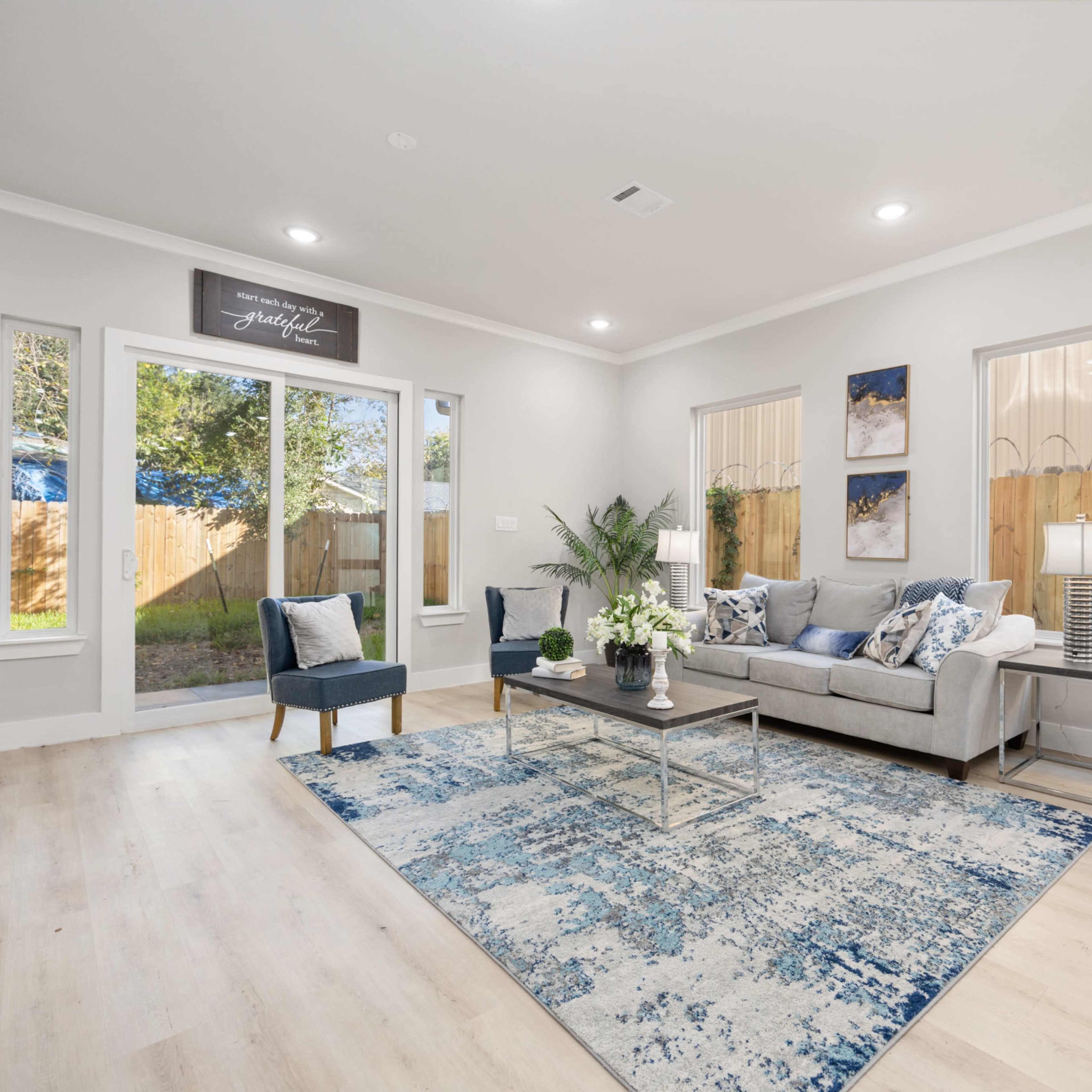 The living room features a gray sofa, a coffee table, and large windows that overlook a backyard, all set on a light-colored wooden floor.