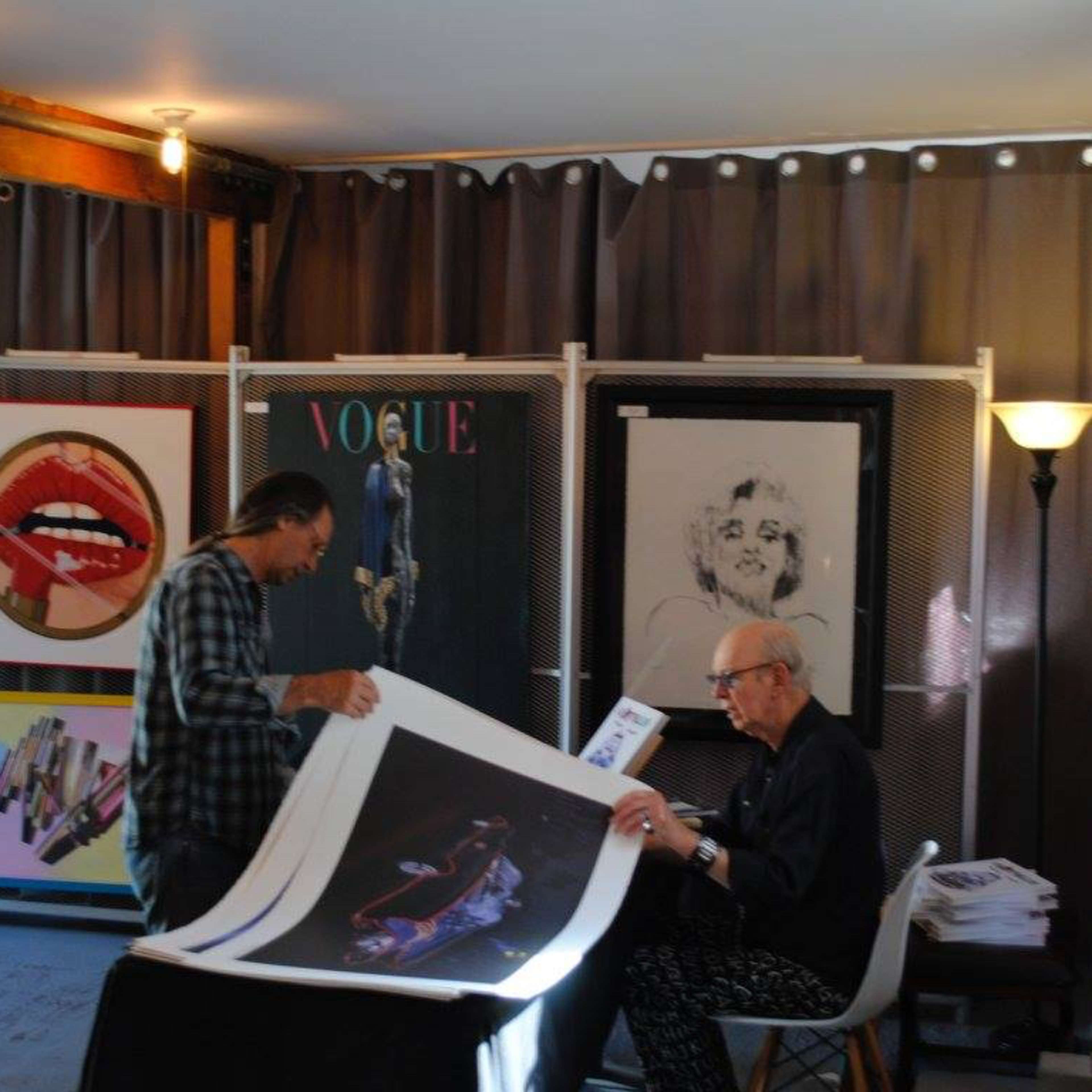 Two men are examining art prints in a studio surrounded by large framed artworks on display.