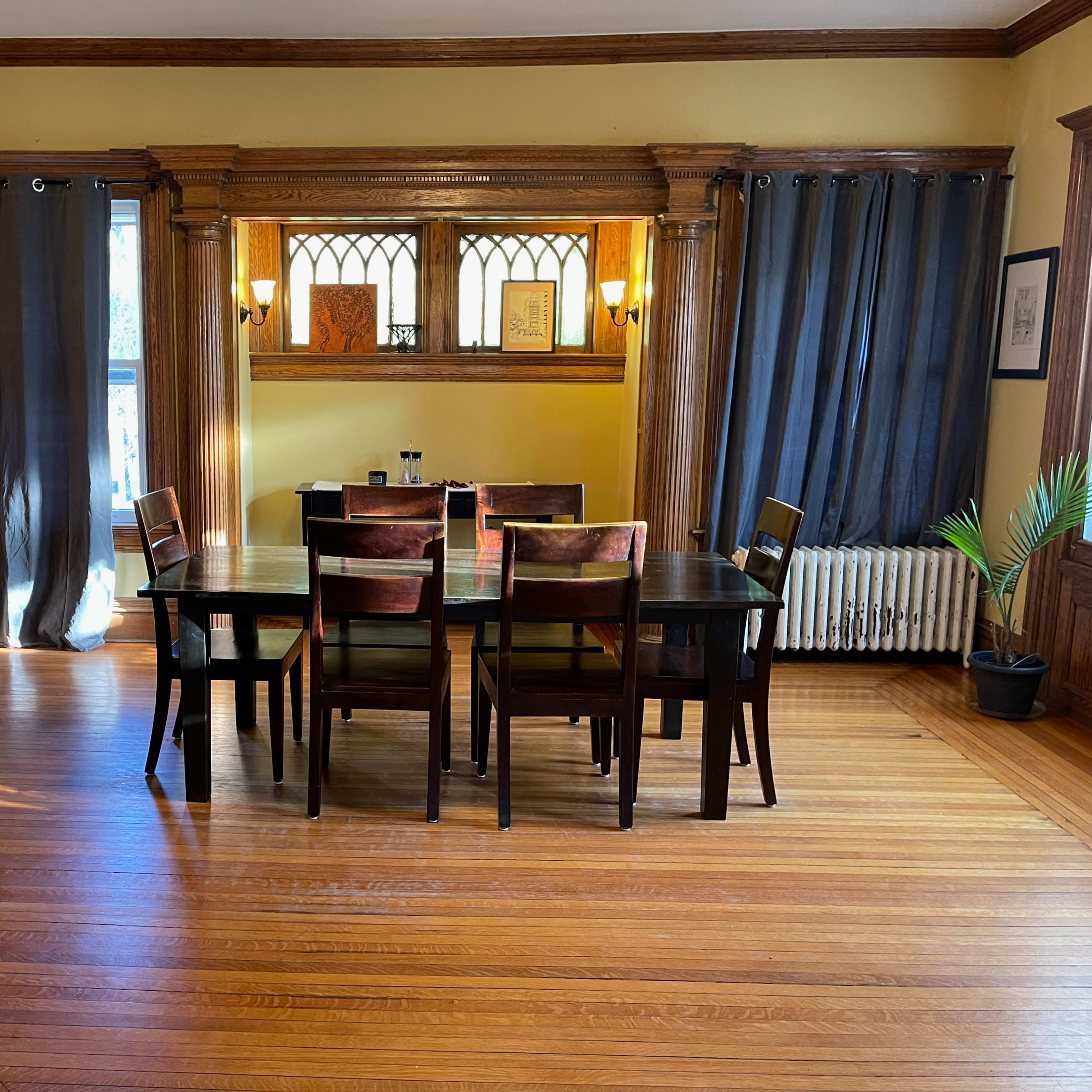 A wooden dining table surrounded by chairs is set in a sunlit room with large windows and dark curtains.