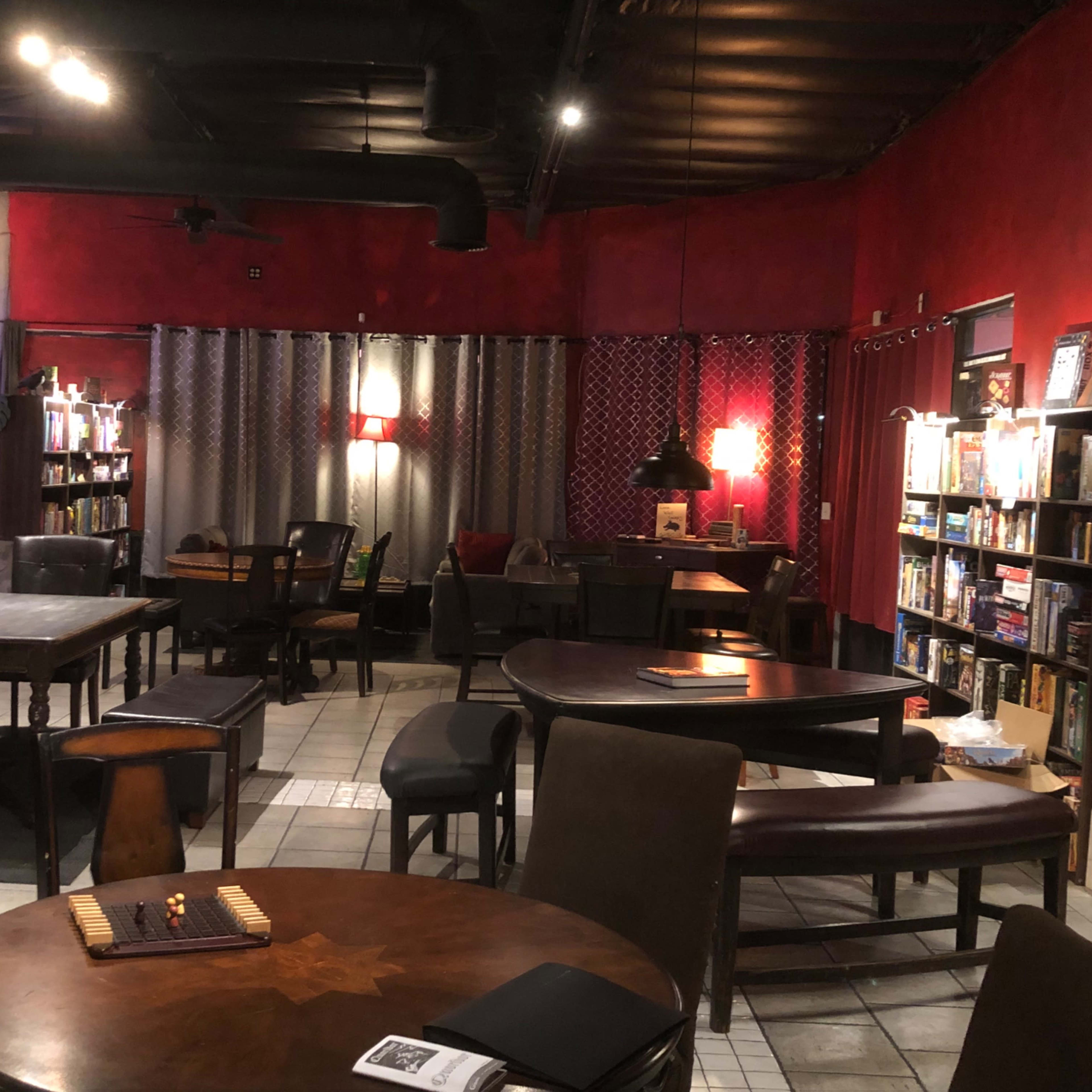 A cozy space with various tables and chairs, bookshelves filled with books, and red walls illuminated by warm lighting.