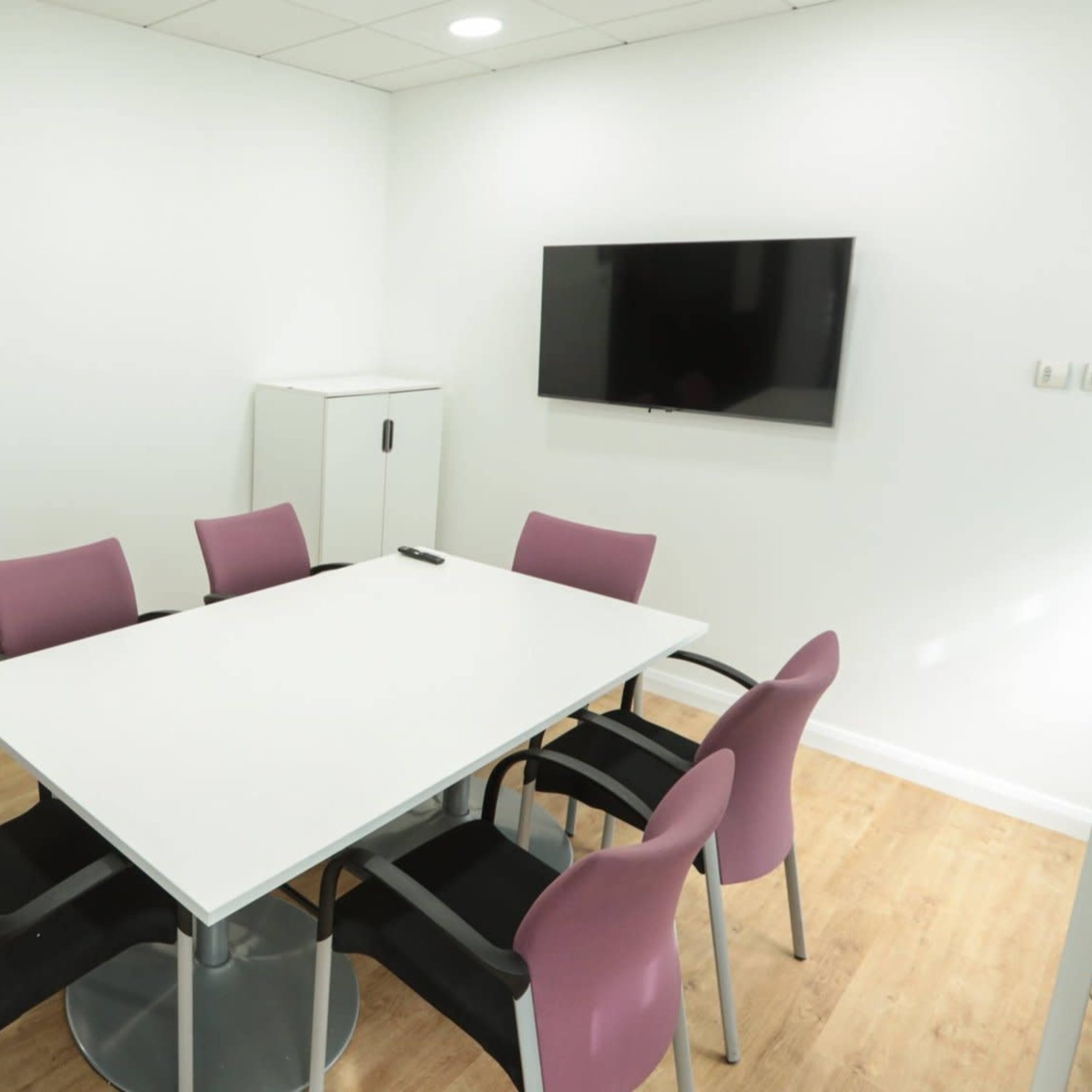 A modern meeting room features a white table surrounded by six chairs, a wall-mounted TV, and a white cabinet.