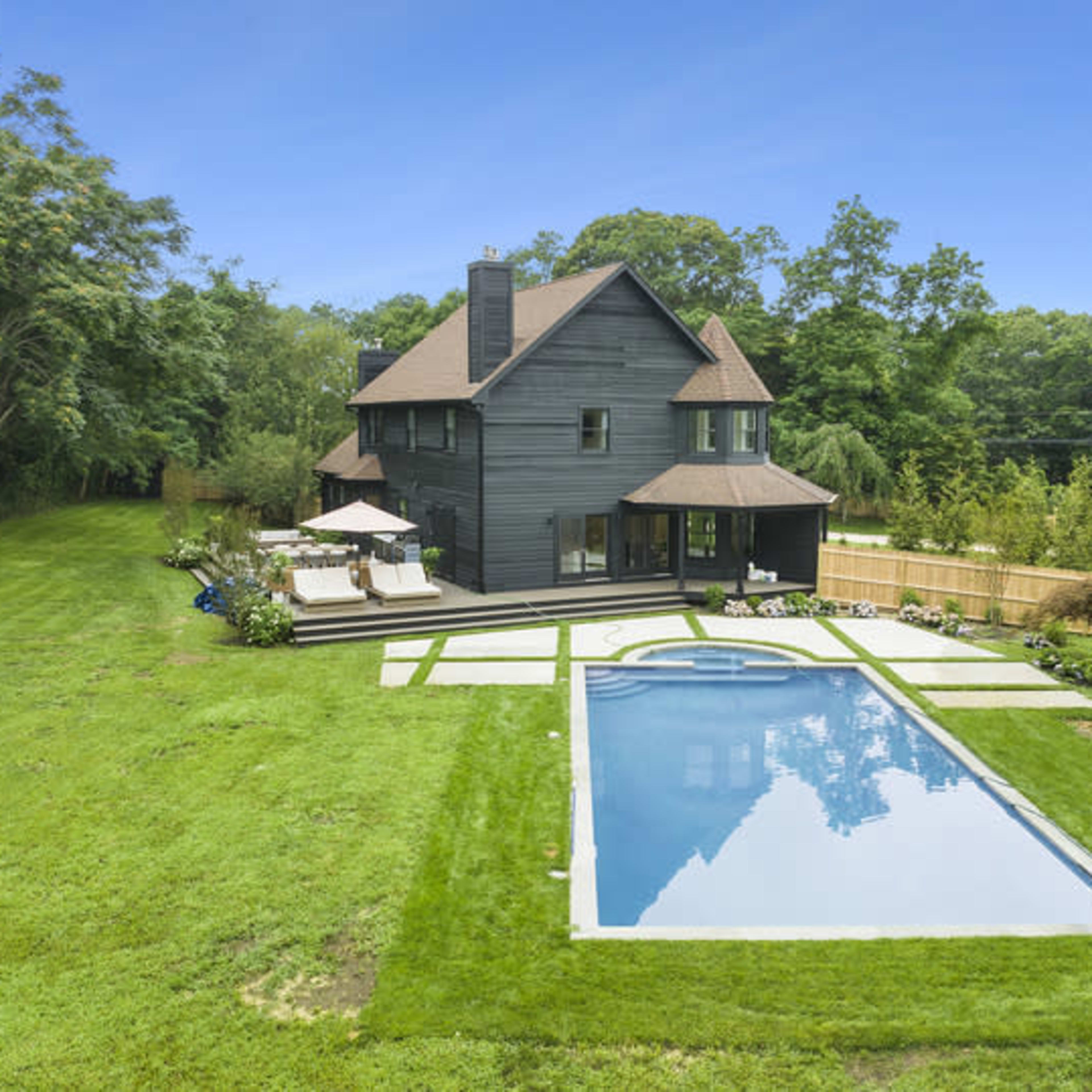 A modern black house is situated near a rectangular swimming pool, surrounded by grass and landscaping.