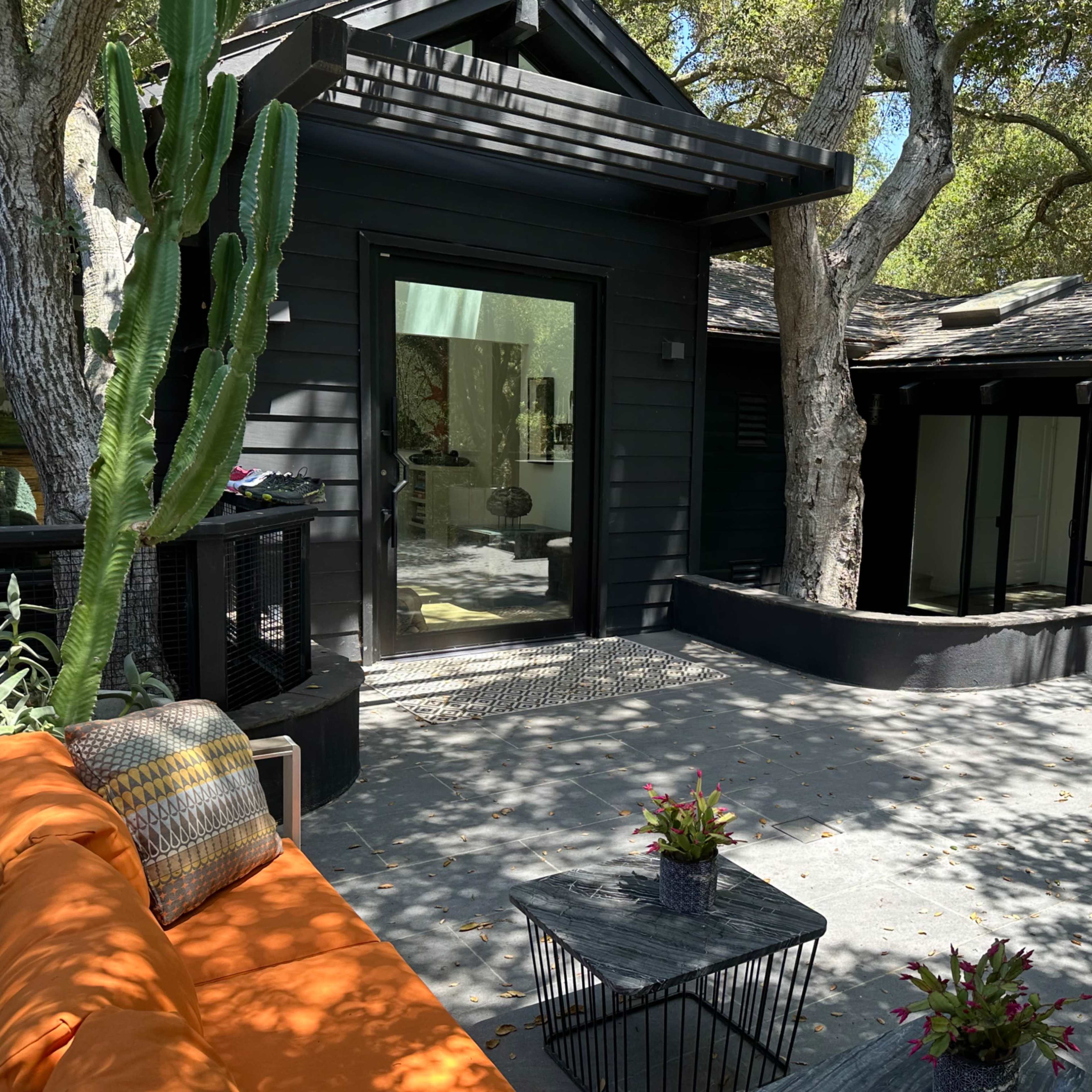 A modern black house surrounded by trees, featuring a patio with an orange sofa and a small table.