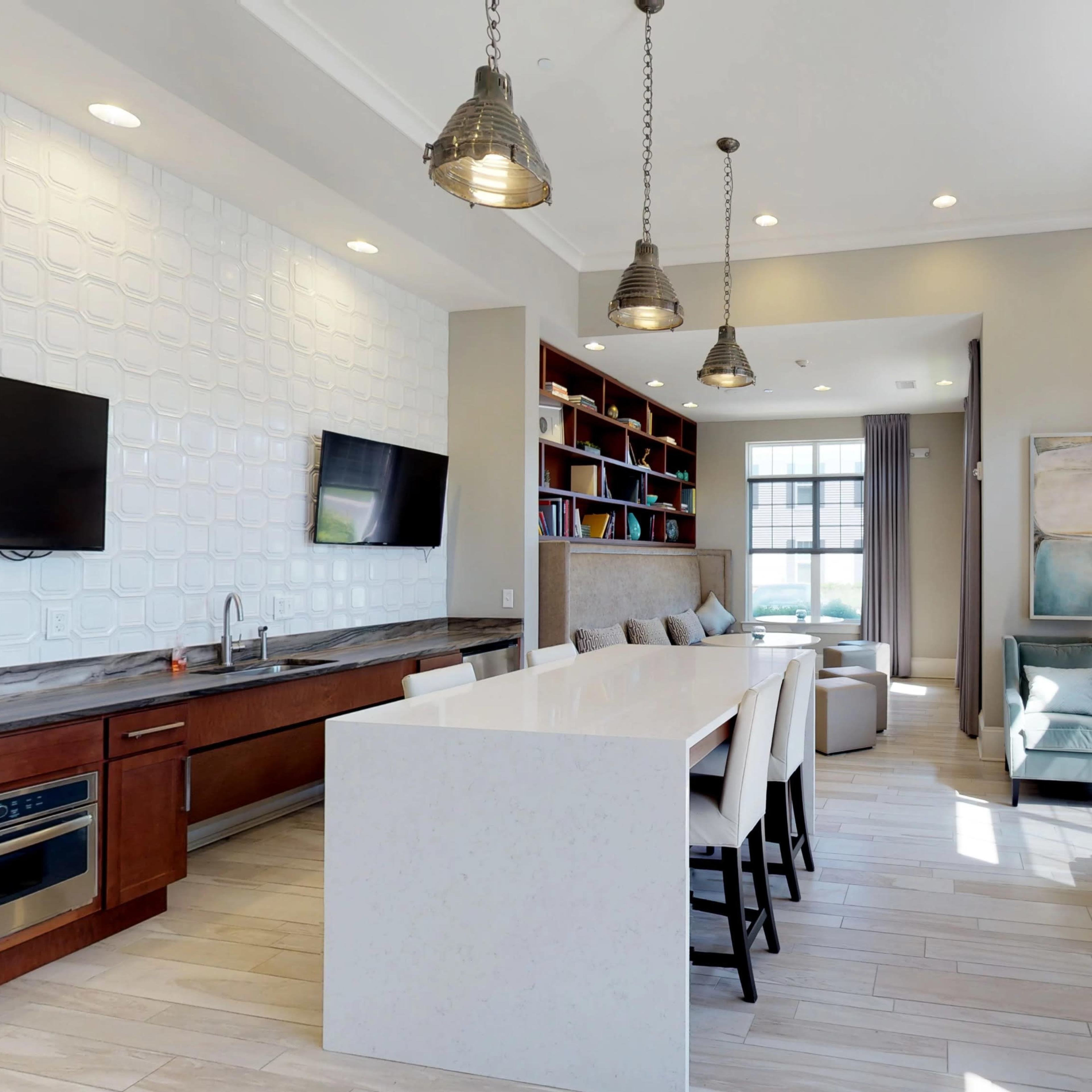 A modern interior space featuring a kitchen area with a large white countertop, seating, bookshelf, and a cozy seating arrangement near windows.