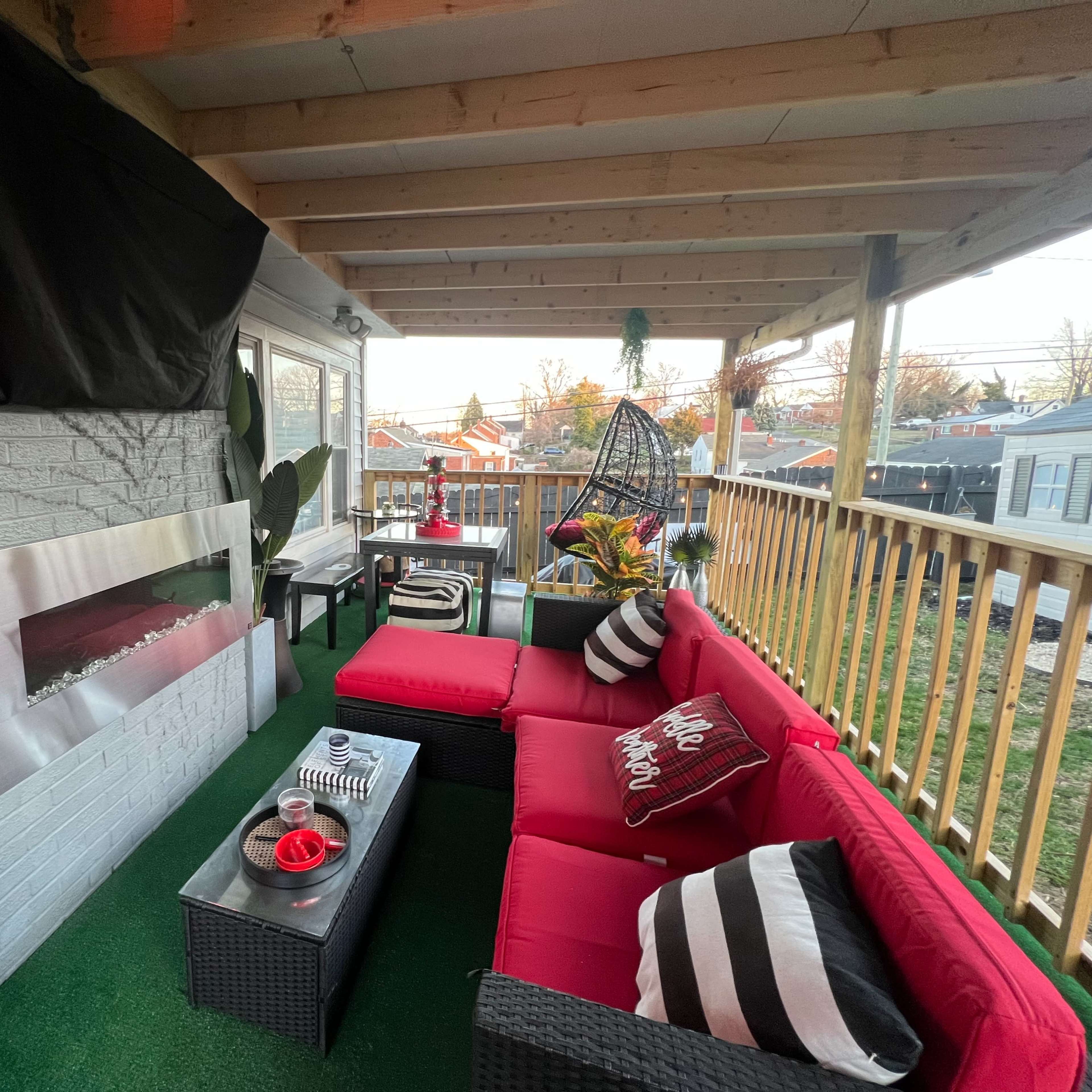 A covered outdoor seating area featuring red couches, a glass coffee table, and a wall-mounted fireplace.