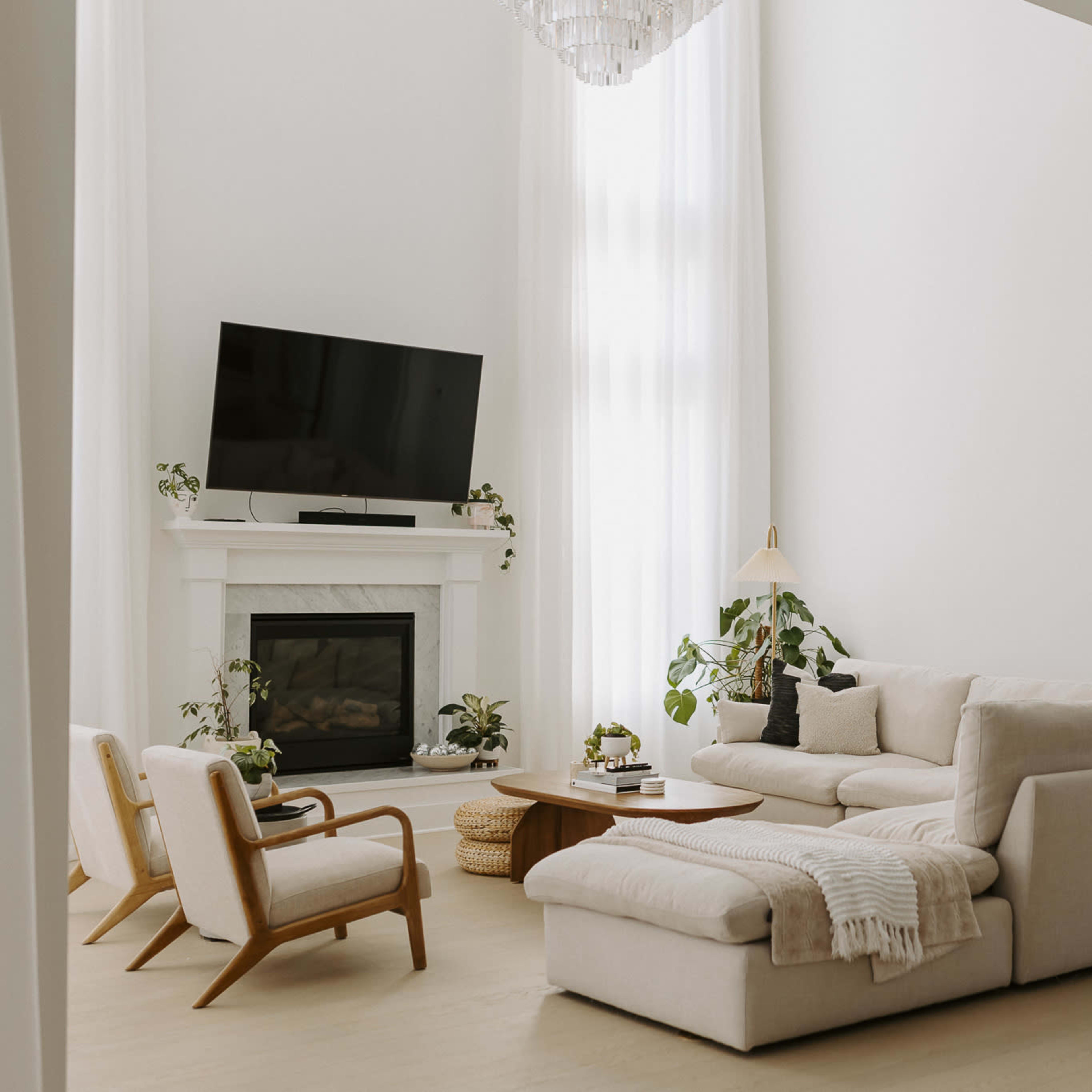 A spacious living room features a light-colored sectional sofa, a wooden coffee table, and a television mounted above a white fireplace, with large windows allowing natural light to fill the space.