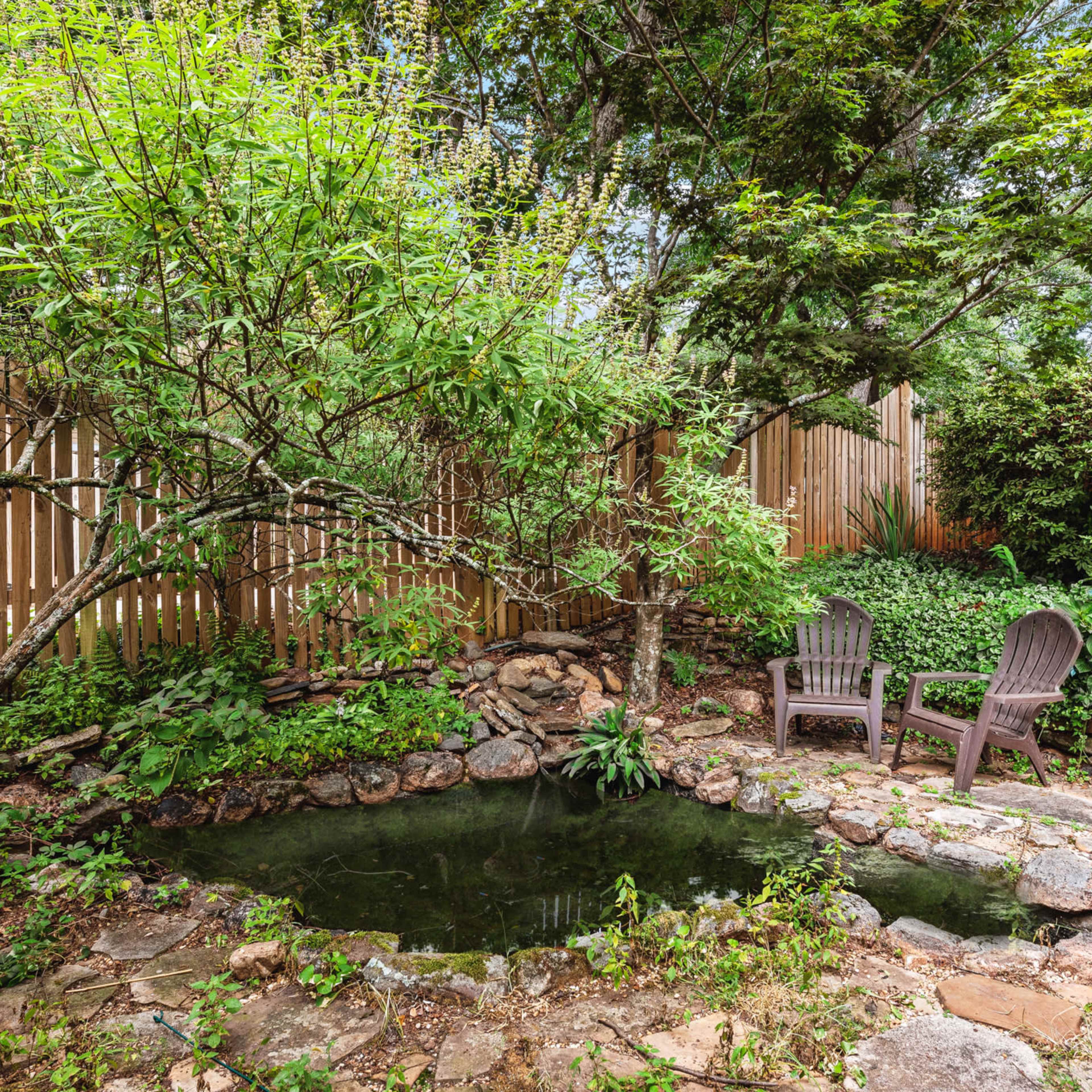 A quiet garden area features a small pond surrounded by rocks and two wooden chairs nestled among greenery.