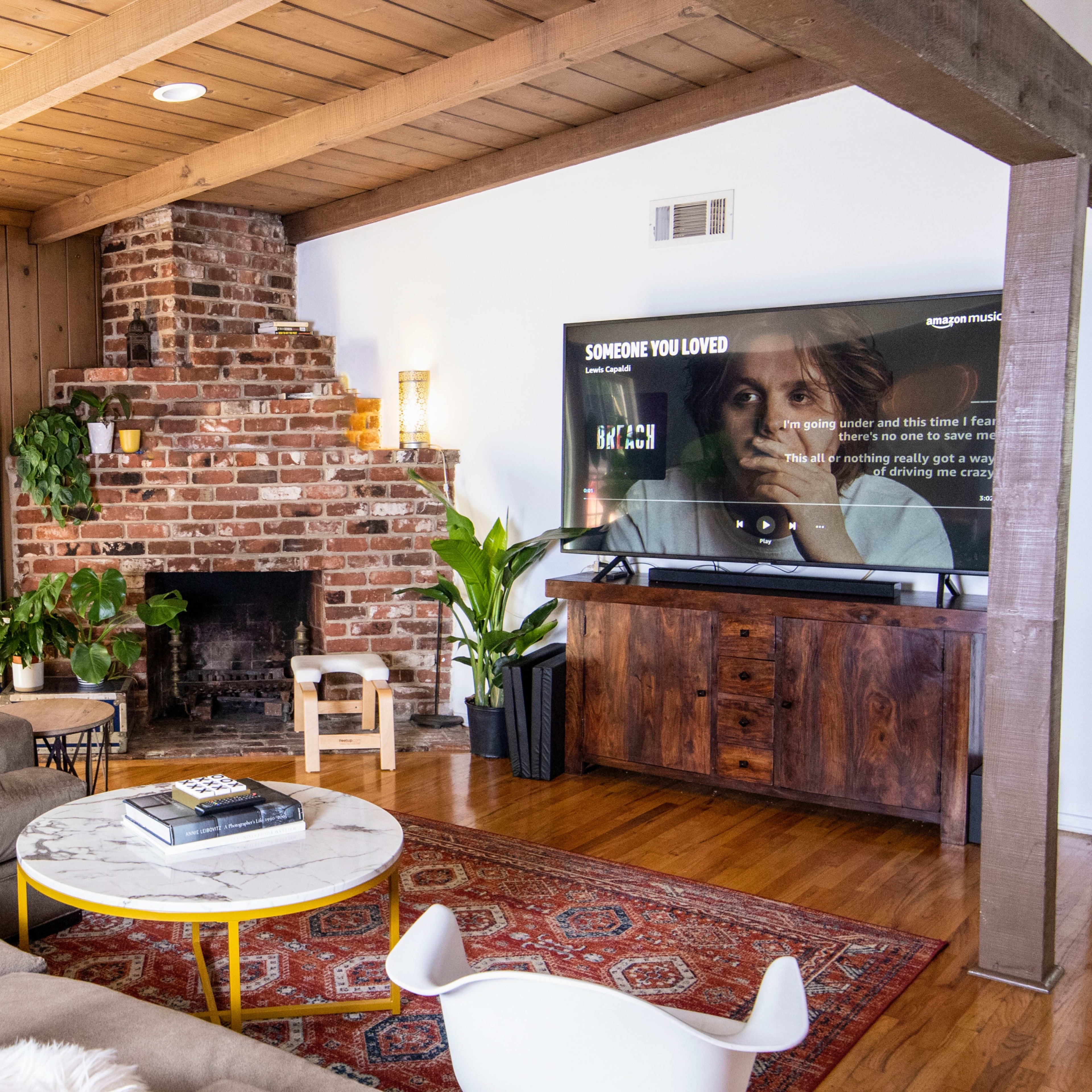A cozy living room features a stone fireplace, a large television displaying a streaming service, and a sofa arranged around a coffee table with plants and decorative items.