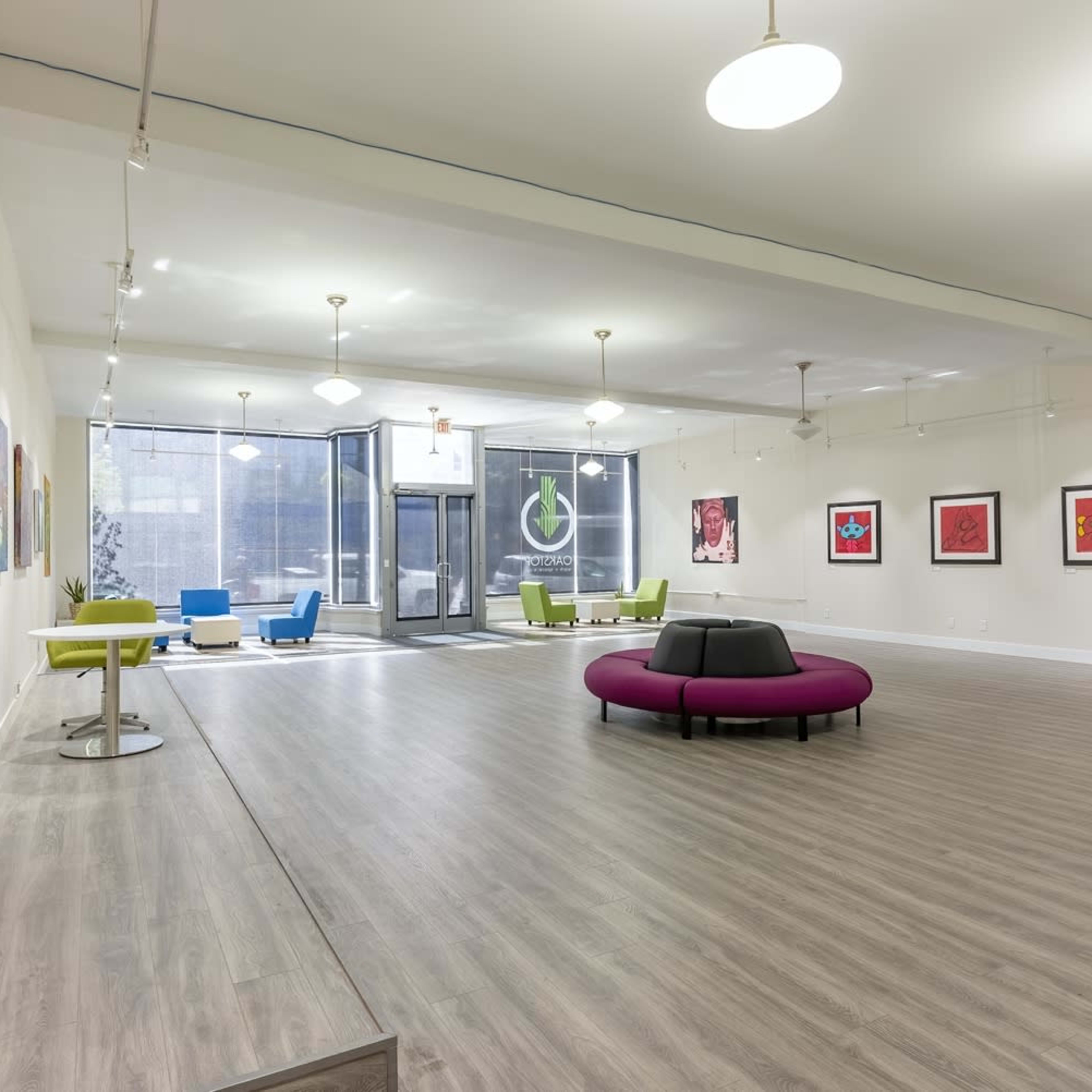 A spacious art gallery with colorful paintings on white walls, modern seating in the center, and large windows letting in natural light.