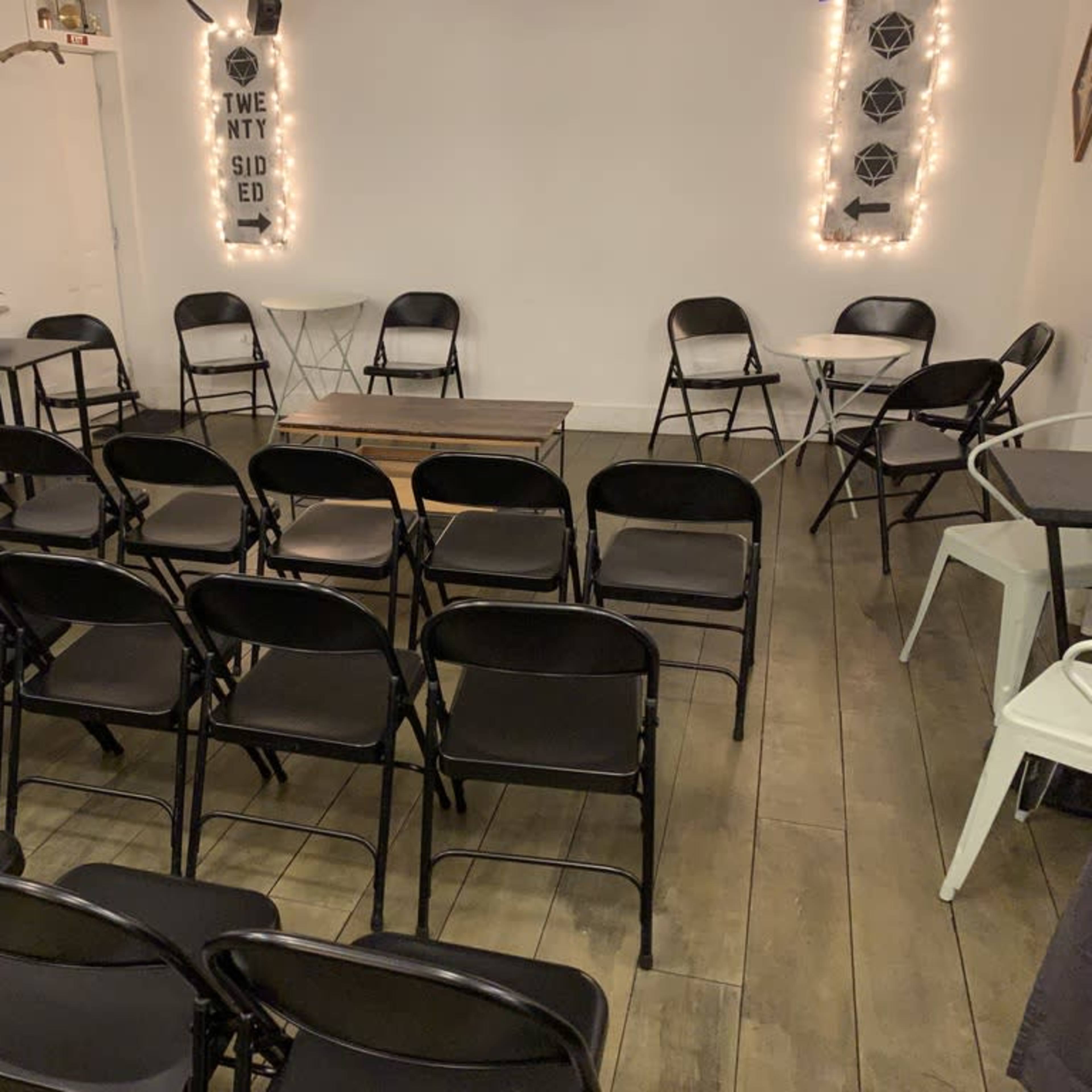 A room with black folding chairs arranged in rows facing a central table, surrounded by white walls and soft lighting.