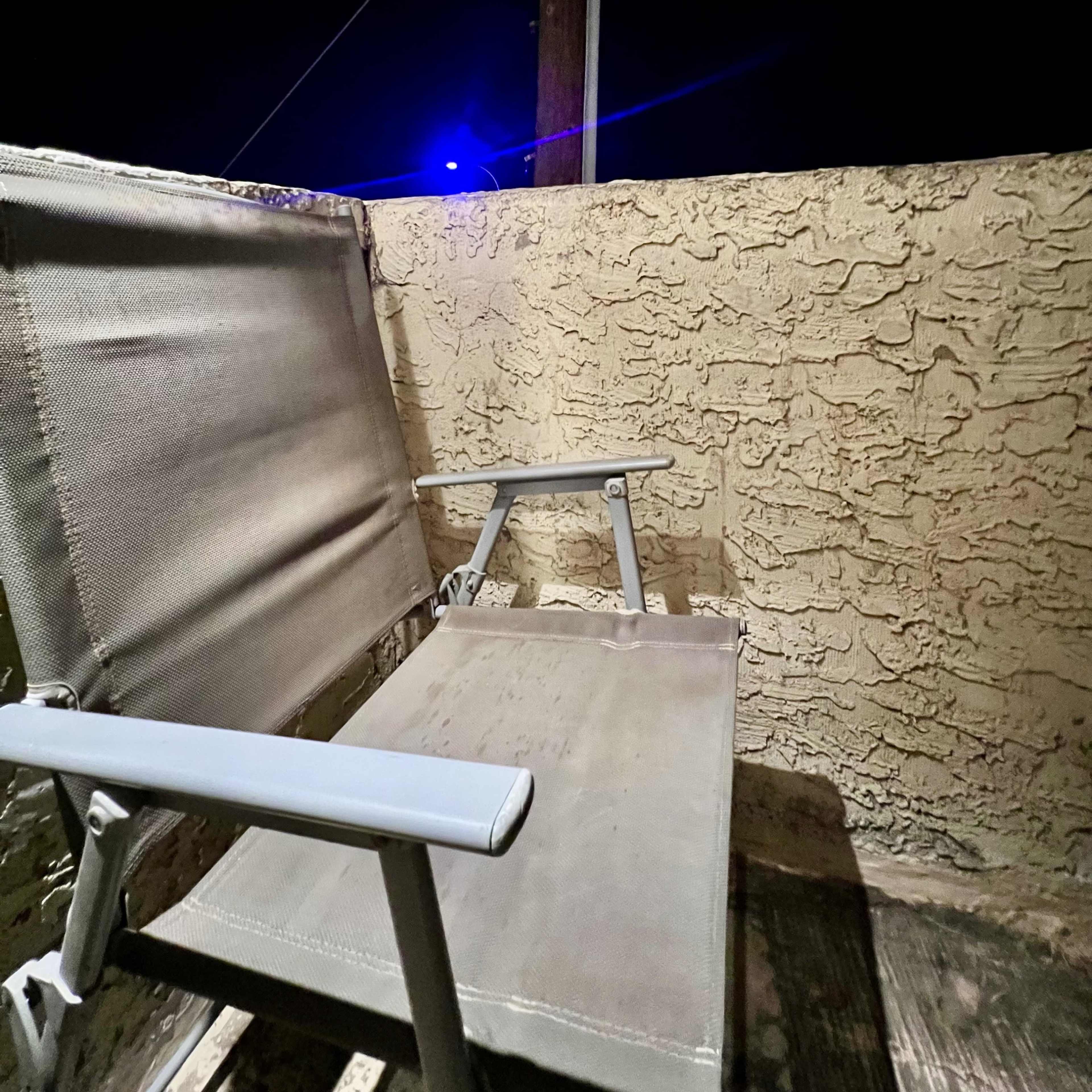 A metal chair sits on a balcony at night, next to a textured wall, with a blue light glowing in the background.