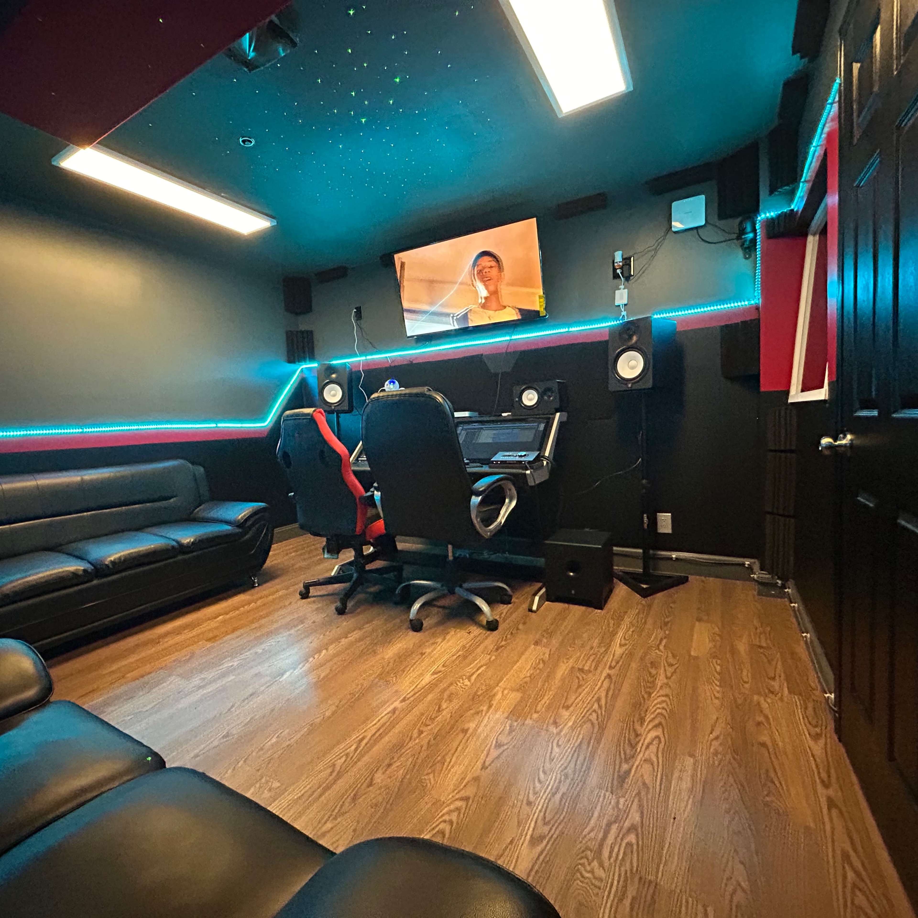 A small recording studio featuring a couch, two office chairs, a computer workstation, and a screen on the wall.