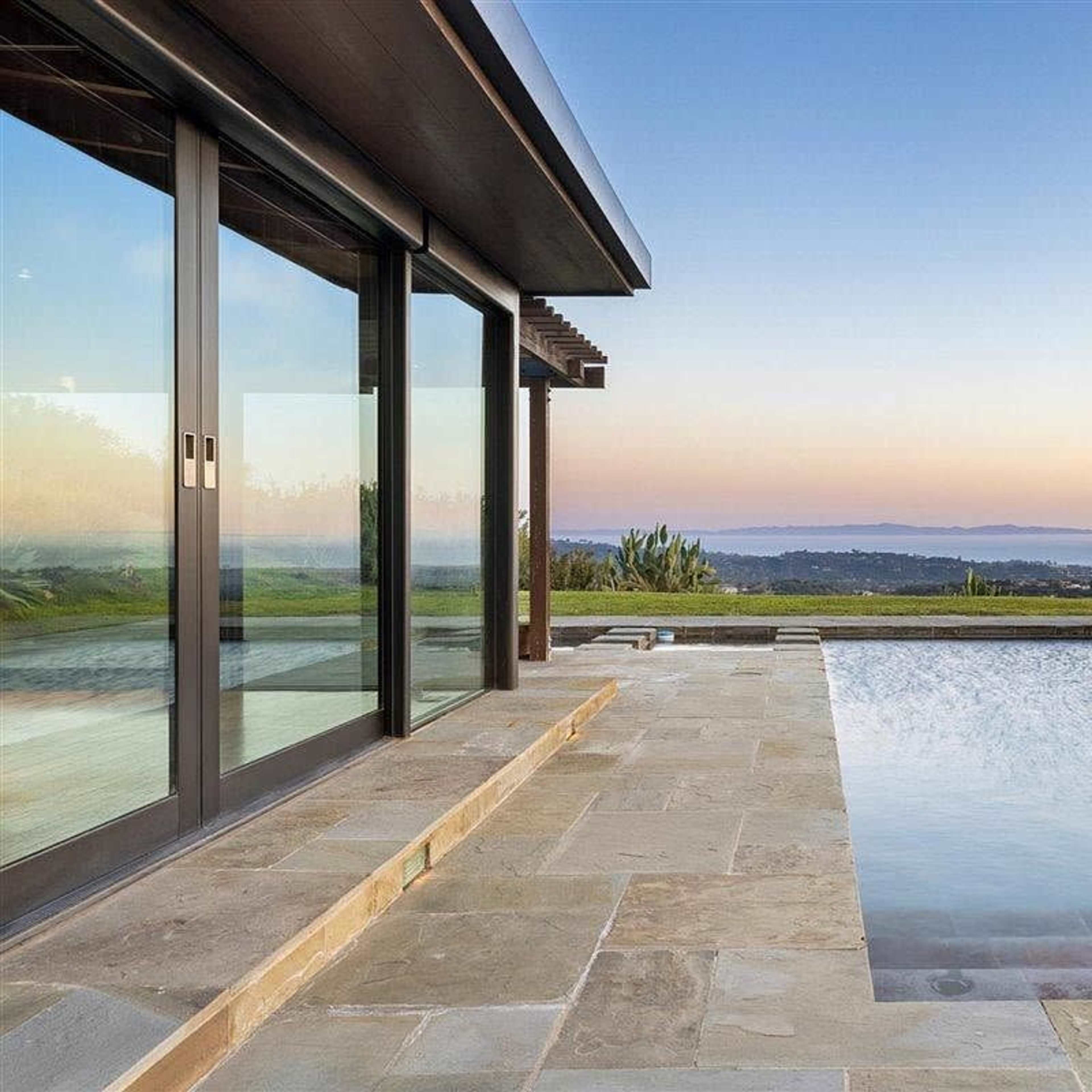 A modern house with large glass doors overlooking a swimming pool and a scenic landscape at sunset.