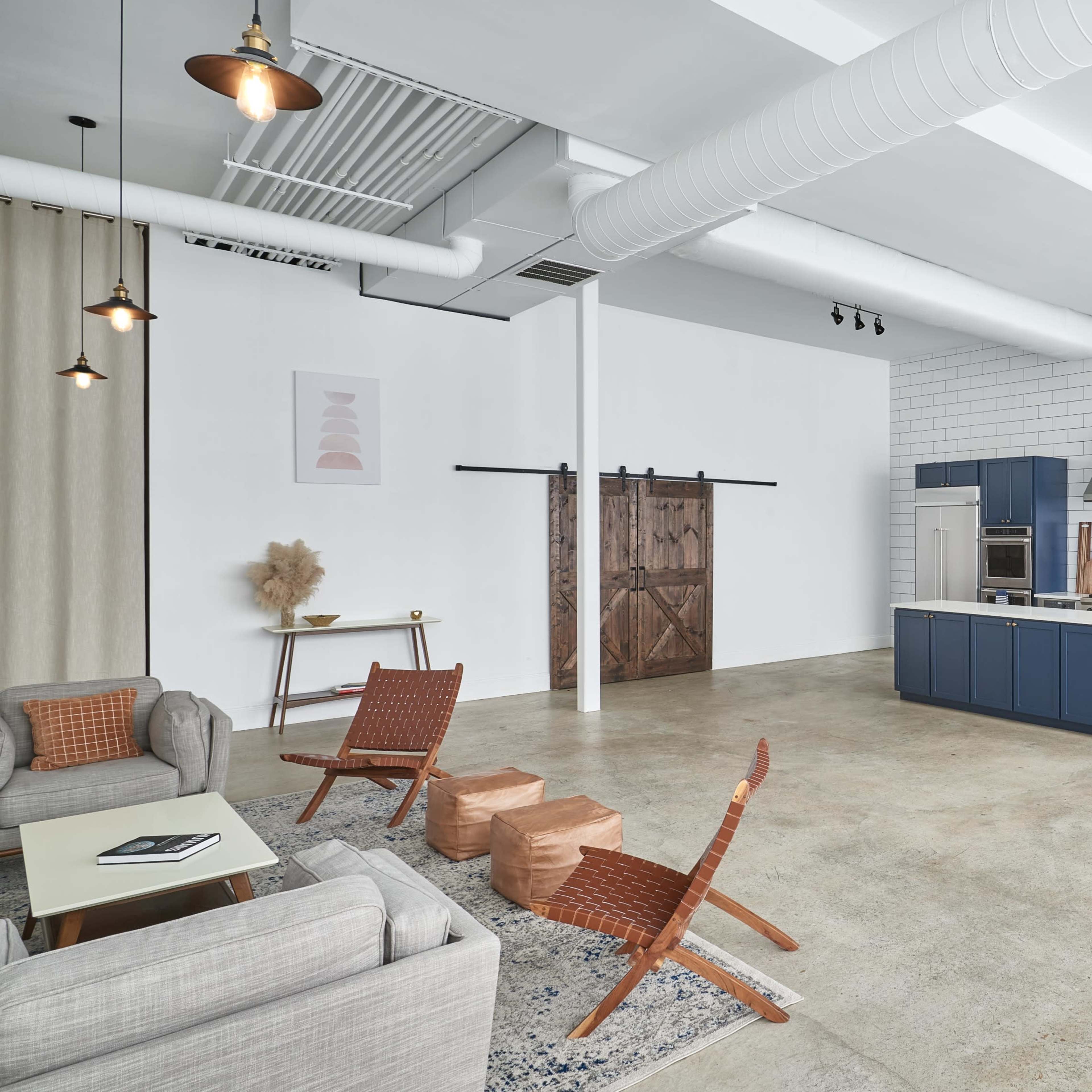 A spacious, modern interior features a kitchen with blue cabinetry, a dining area with a wooden table and blue chairs, and a lounge with gray sofas and woven chairs.