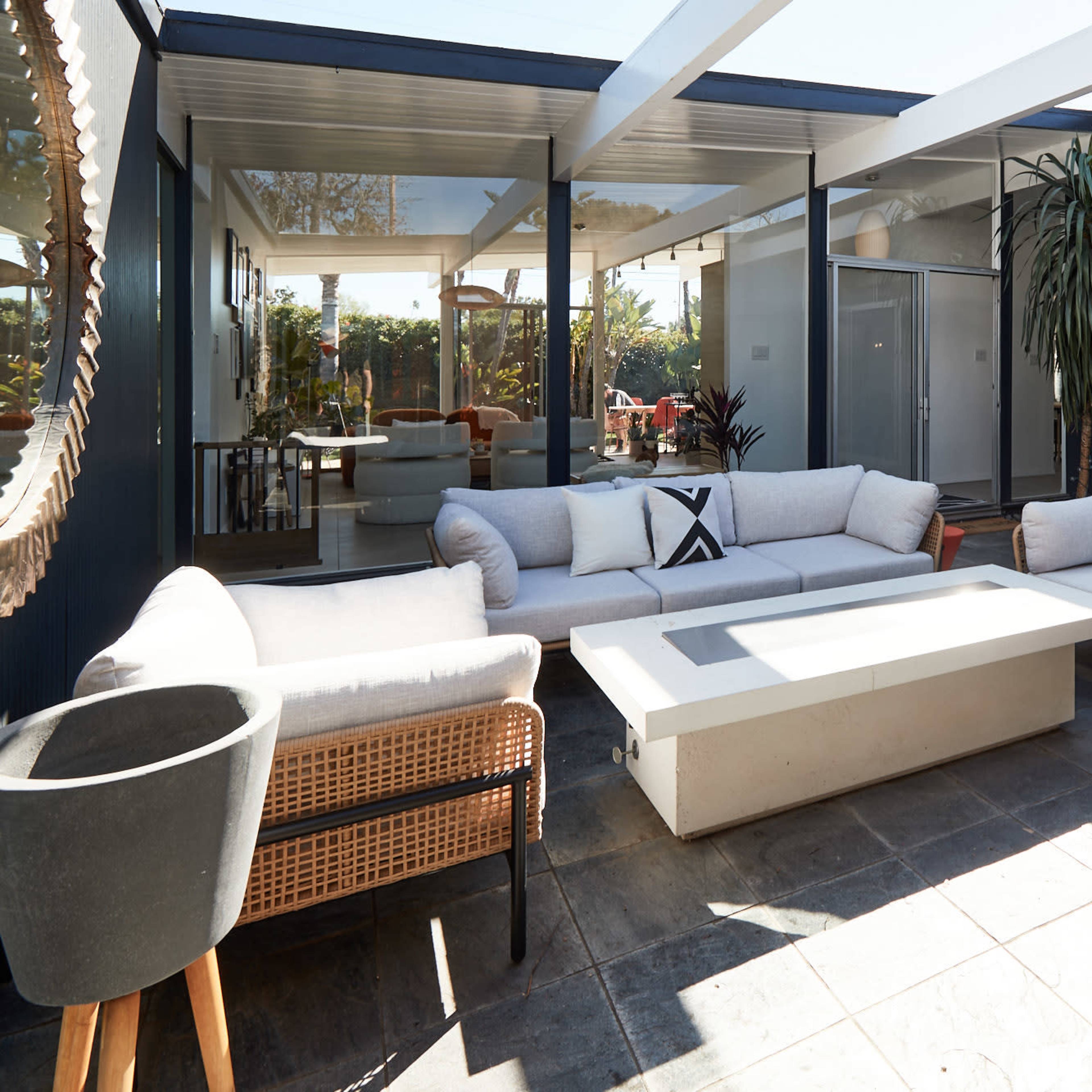 A modern outdoor living space featuring a white sofa set and a rectangular coffee table on a stone patio, surrounded by glass walls and greenery.