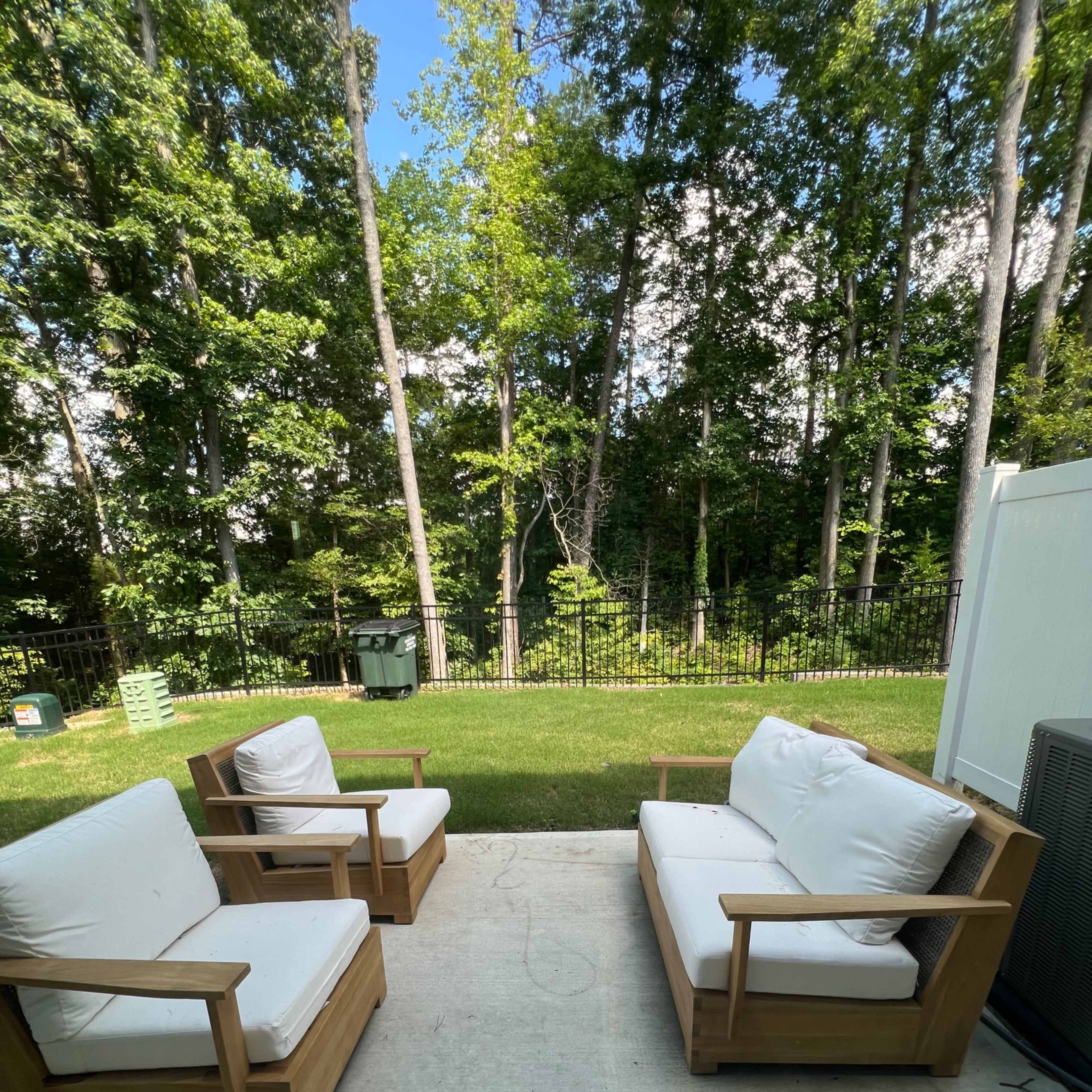 A small patio area with two wooden armchairs and a cushioned sofa facing a green lawn and dense trees in the background.