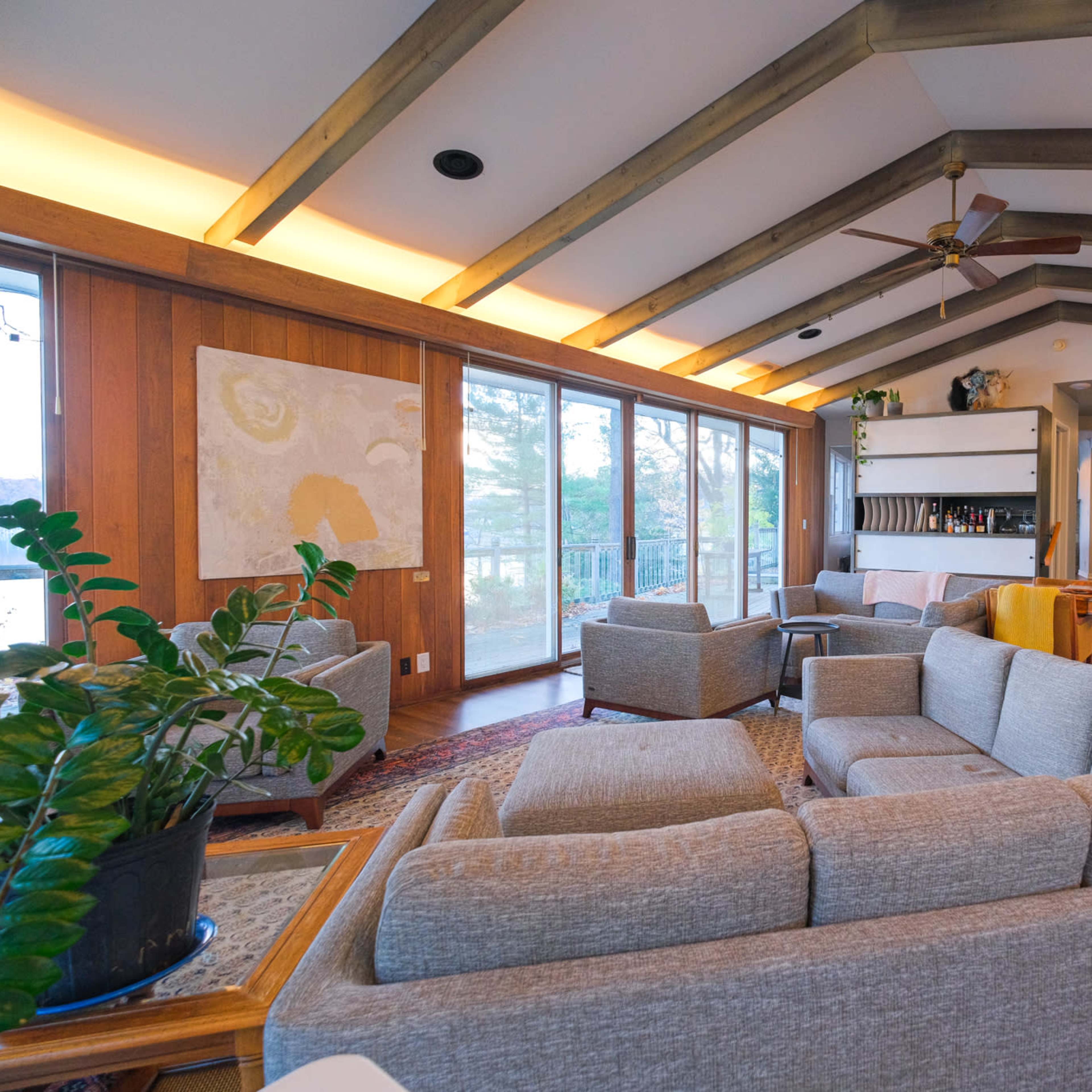 A modern living room with a light gray sectional sofa, wooden beams on the ceiling, and large windows overlooking a natural view.