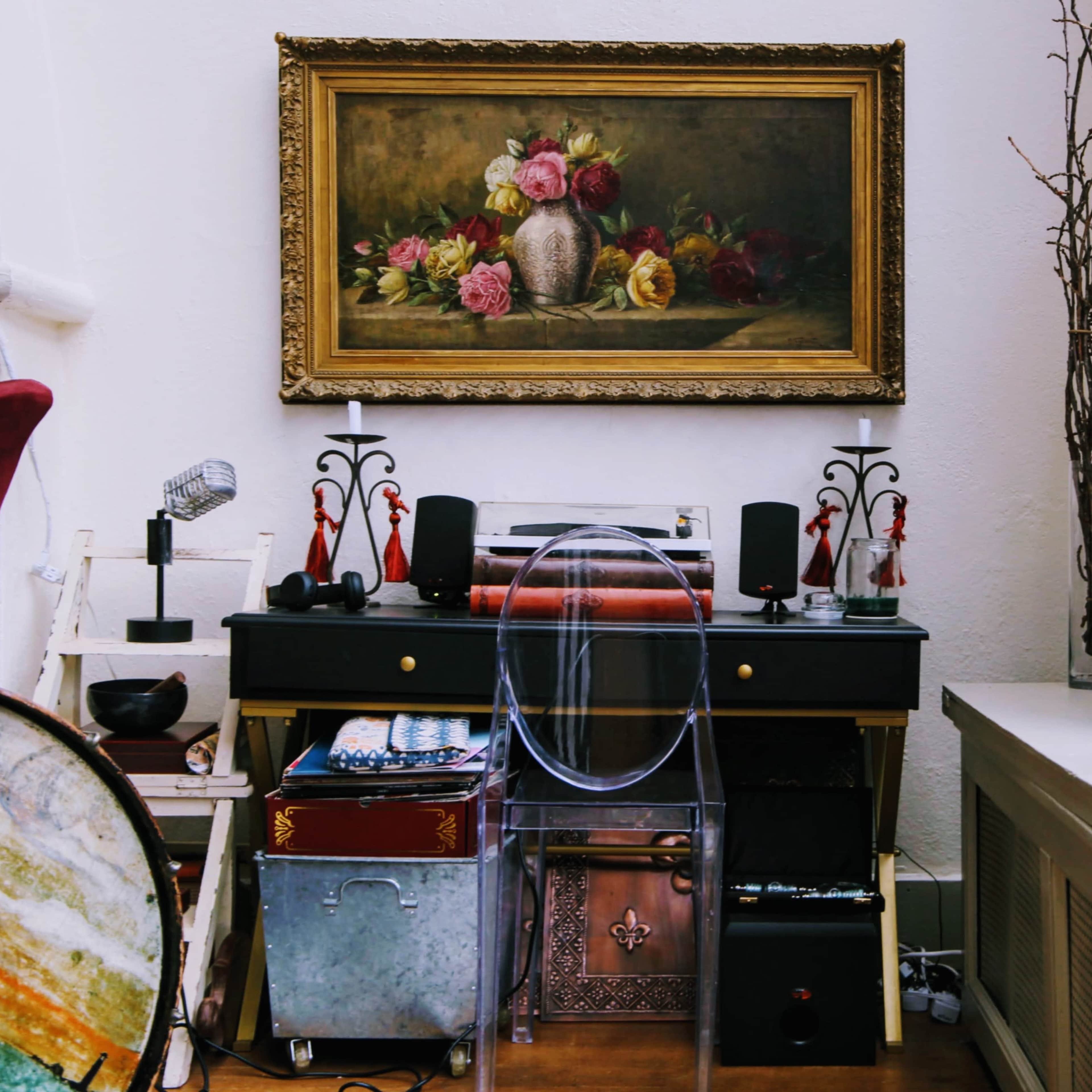 A vintage-style desk with a clear chair is positioned beneath a floral painting, surrounded by decorative elements and storage boxes.
