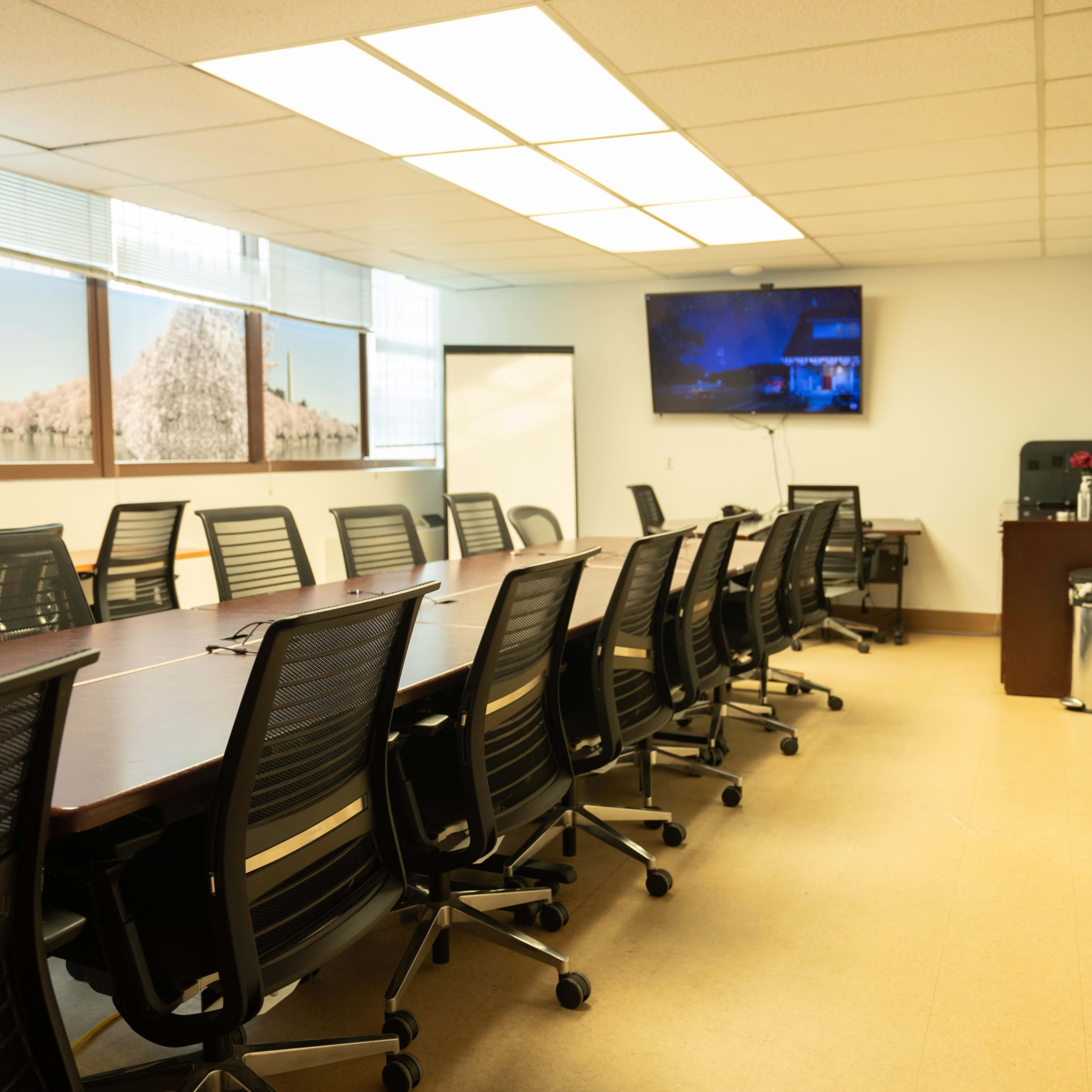 A conference room with a long table and multiple black chairs, featuring a large screen and windows with a view.