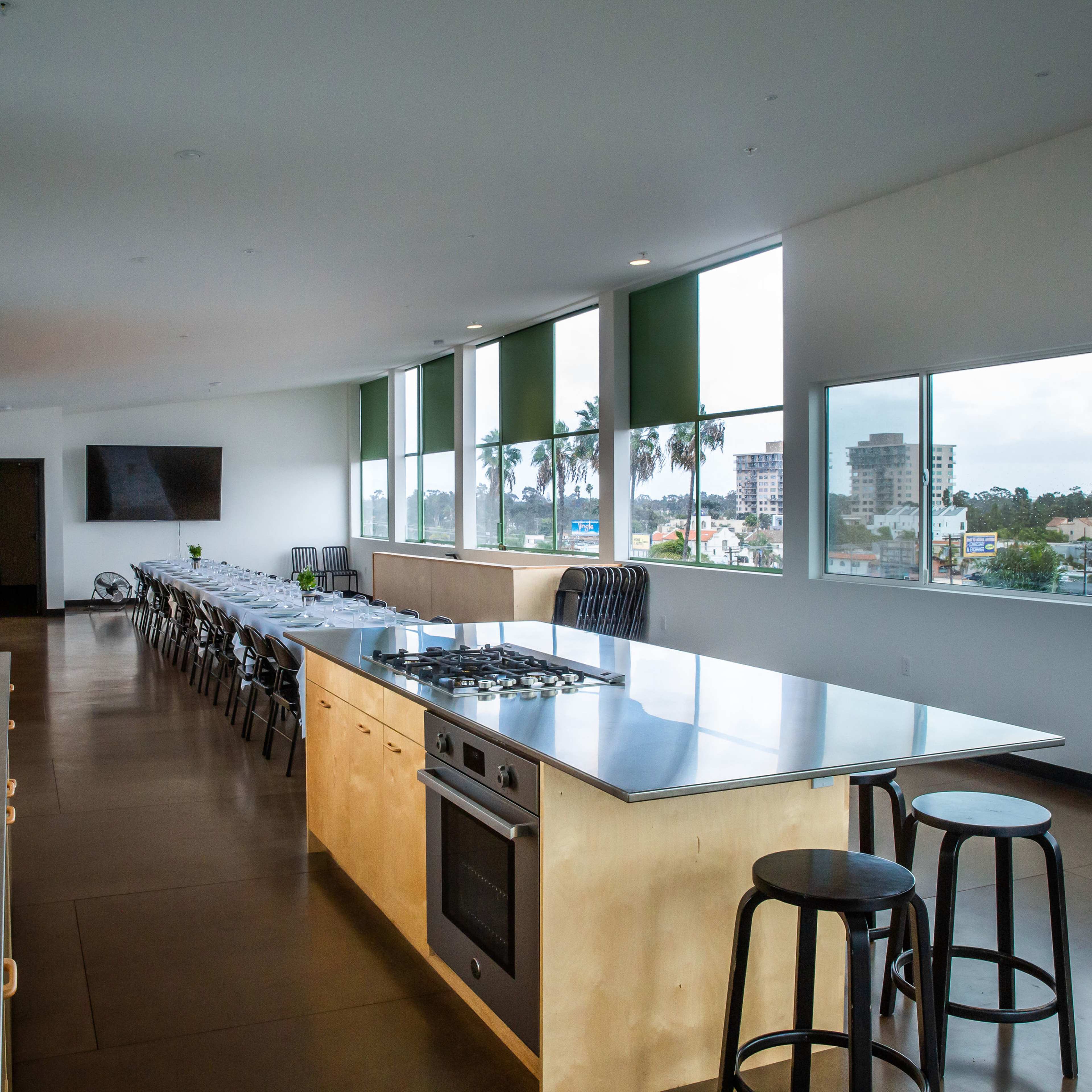 The kitchen features a long table lined with chairs, a stainless steel countertop, and large windows offering a view of the city.