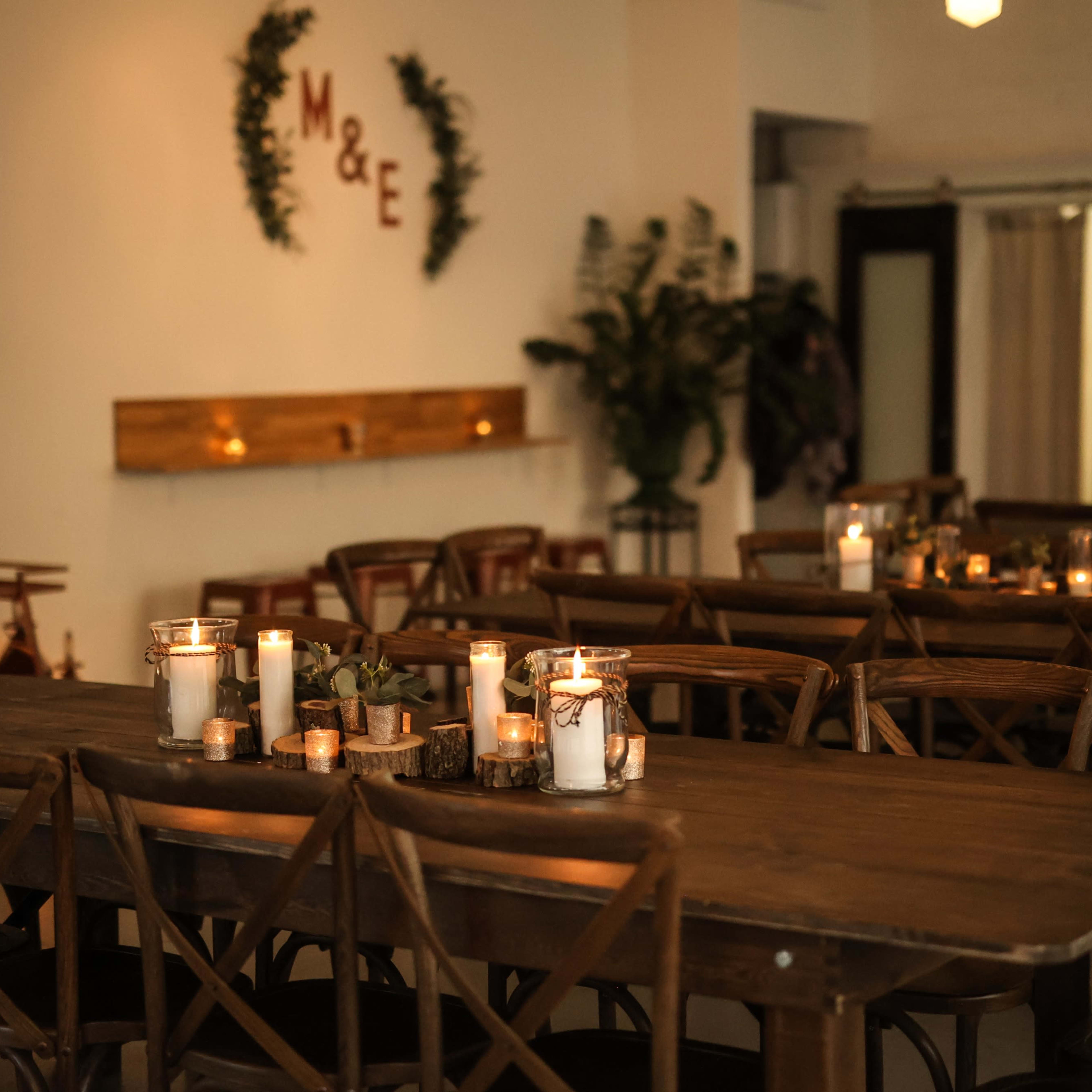 A warmly lit event space with wooden tables arranged for seating, adorned with candles and simple decorations.
