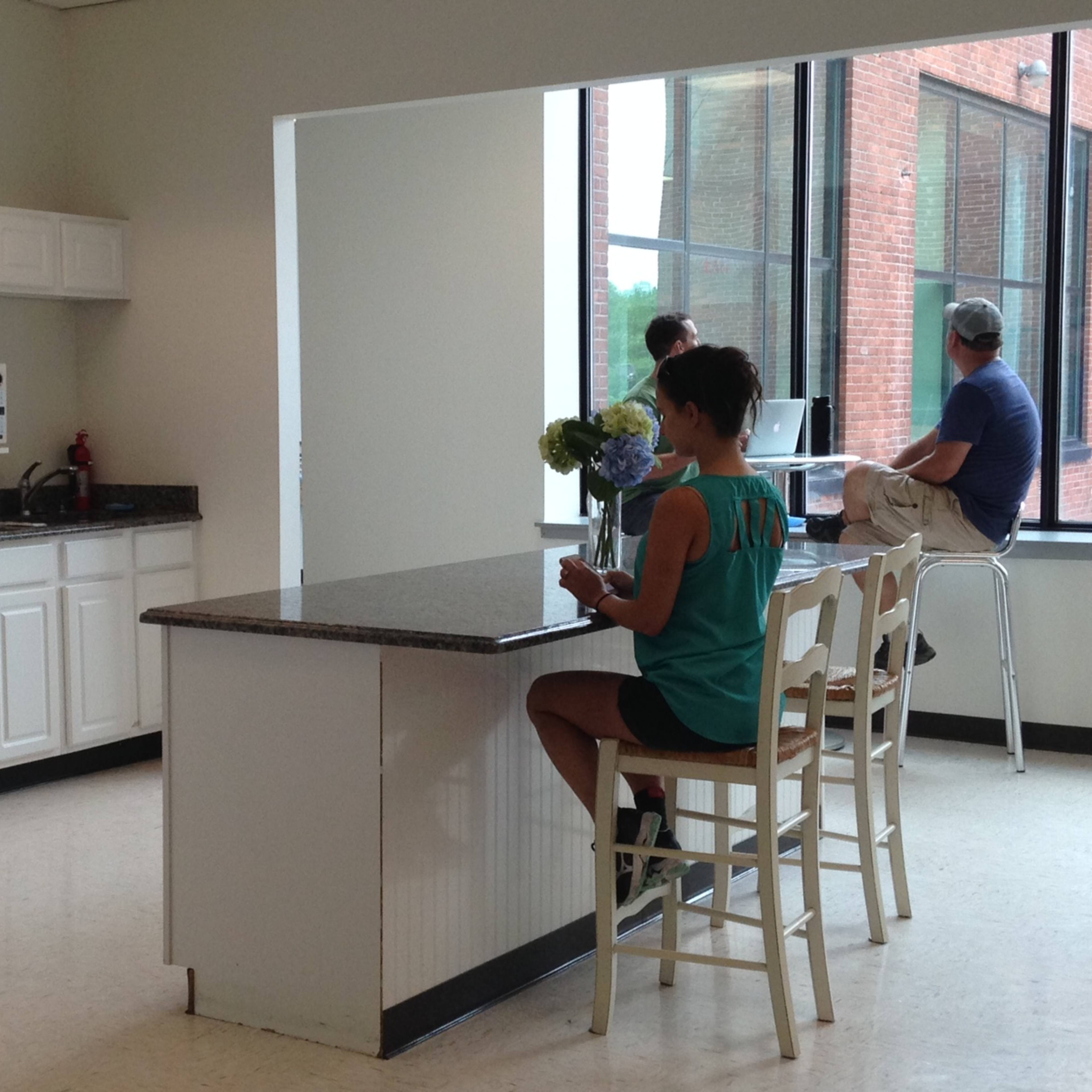 A kitchen area features a person washing dishes, while another sits at a counter and two people work on laptops at a table.