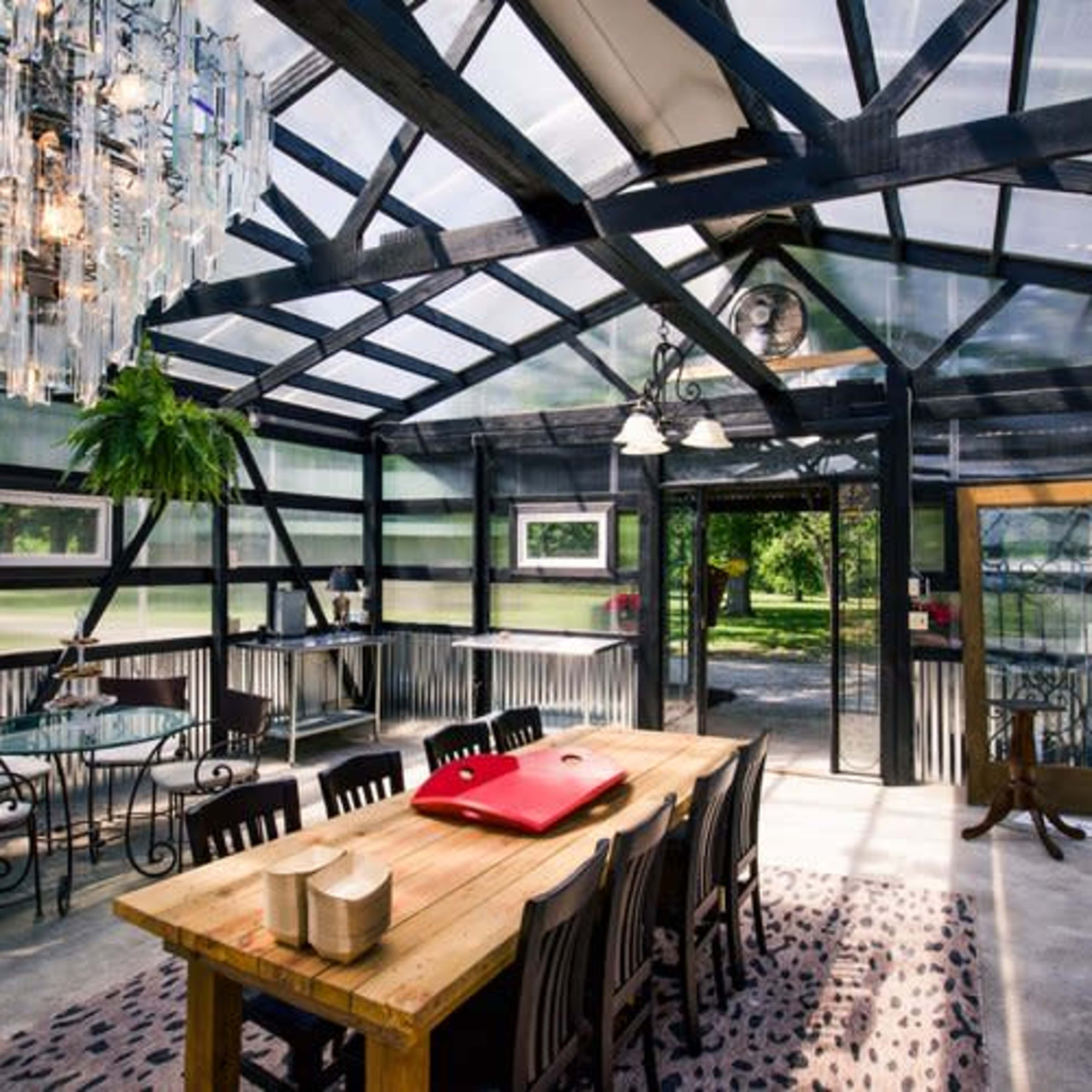 A modern greenhouse dining area featuring a large wooden table, elegant chandelier, and glass walls surrounded by greenery.