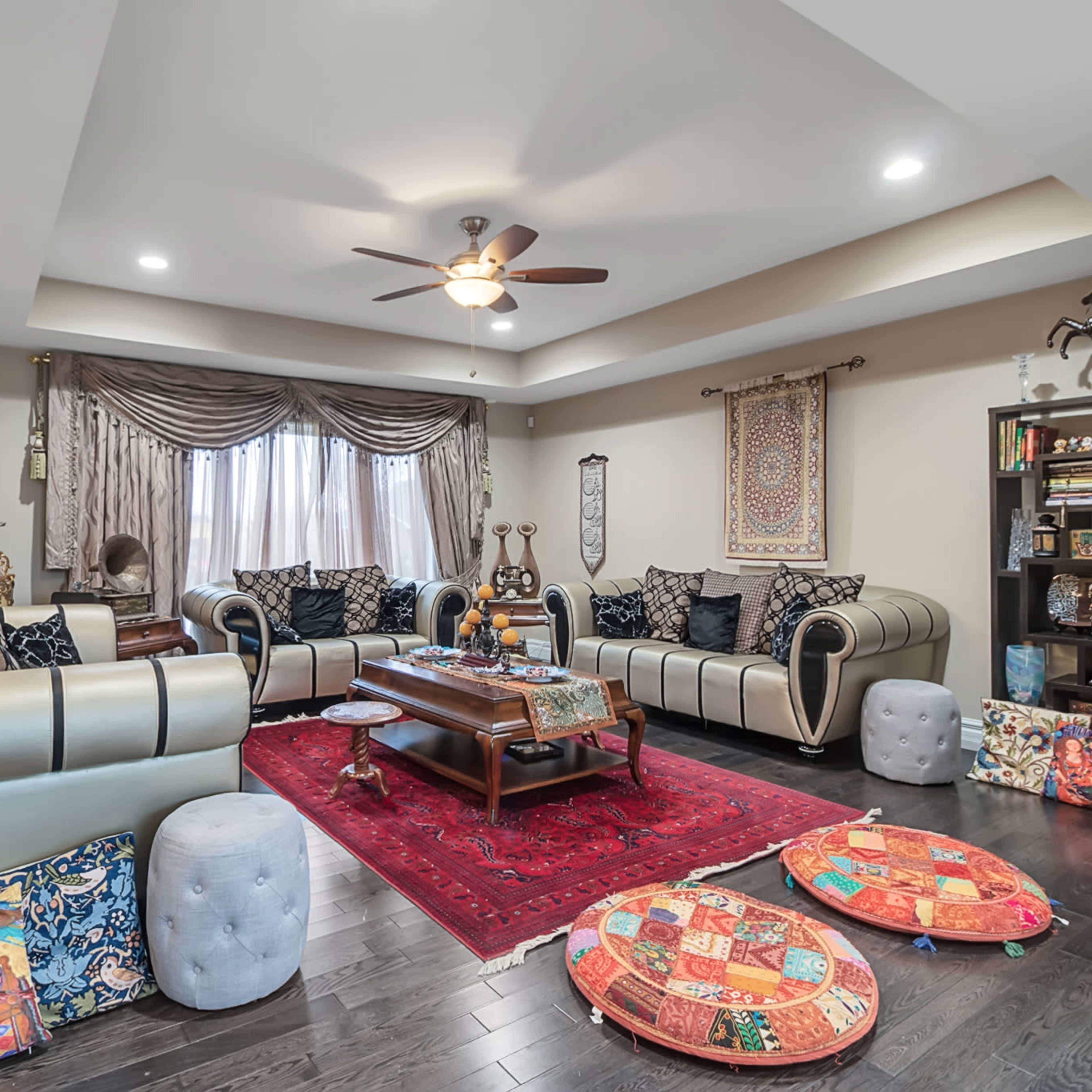 A living room features a large, multi-seated sofa, colorful floor cushions, a wooden coffee table, and decorative items arranged around a red area rug.