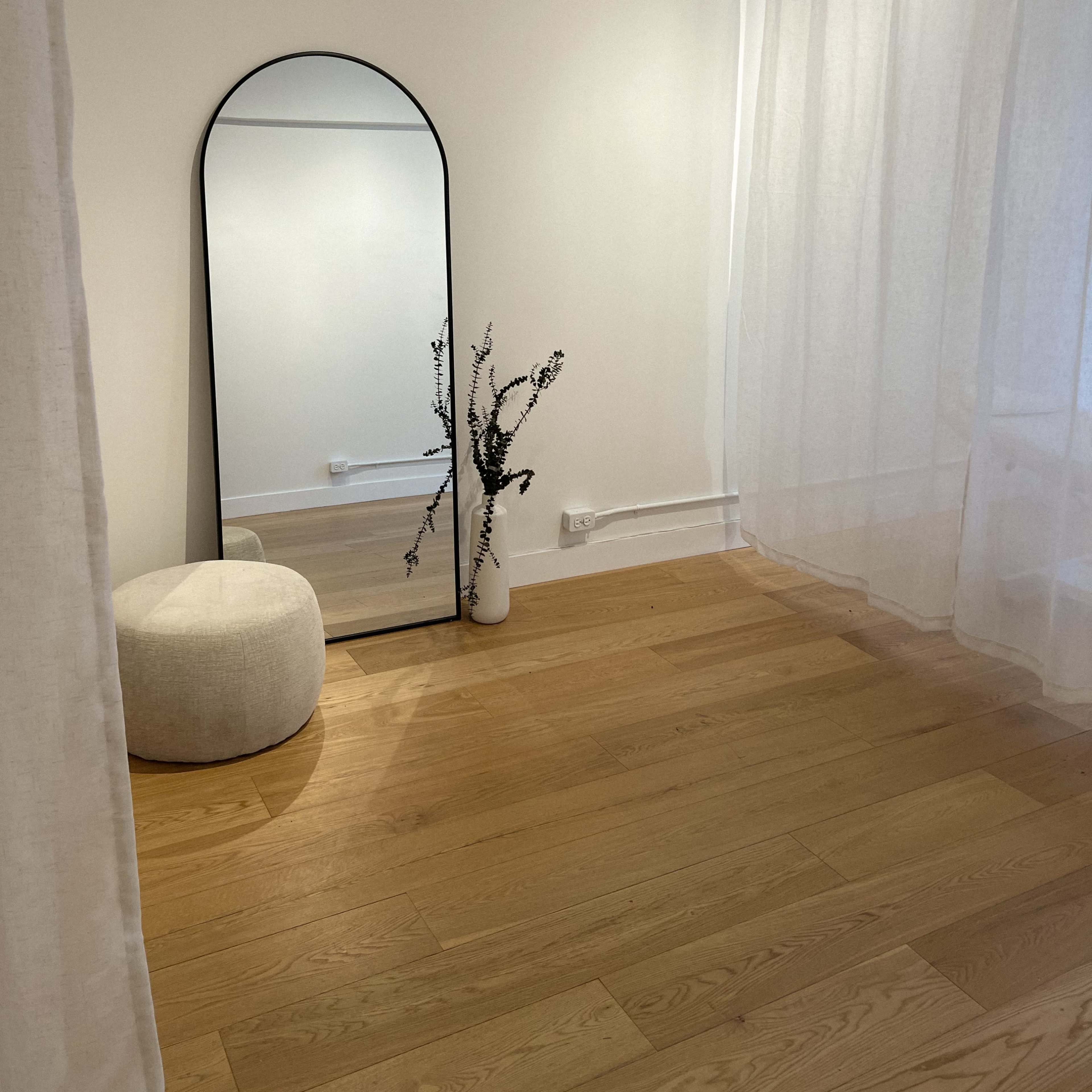 A minimalistic room features a large mirror, a white pouf, and a vase with branches beside sheer curtains and wooden flooring.