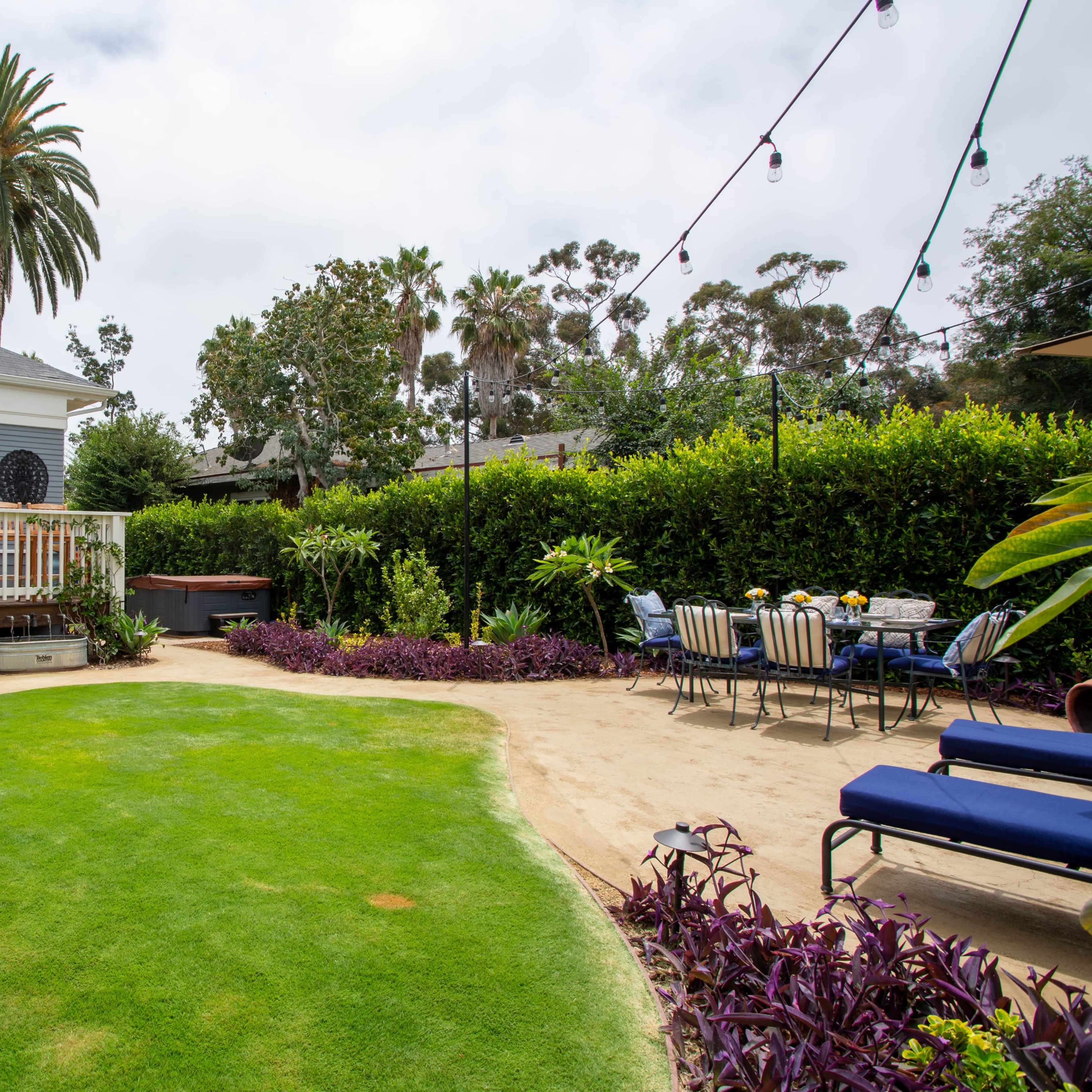 A landscaped backyard features a green lawn, potted plants, a dining table, and a hot tub, surrounded by hedges and palm trees.