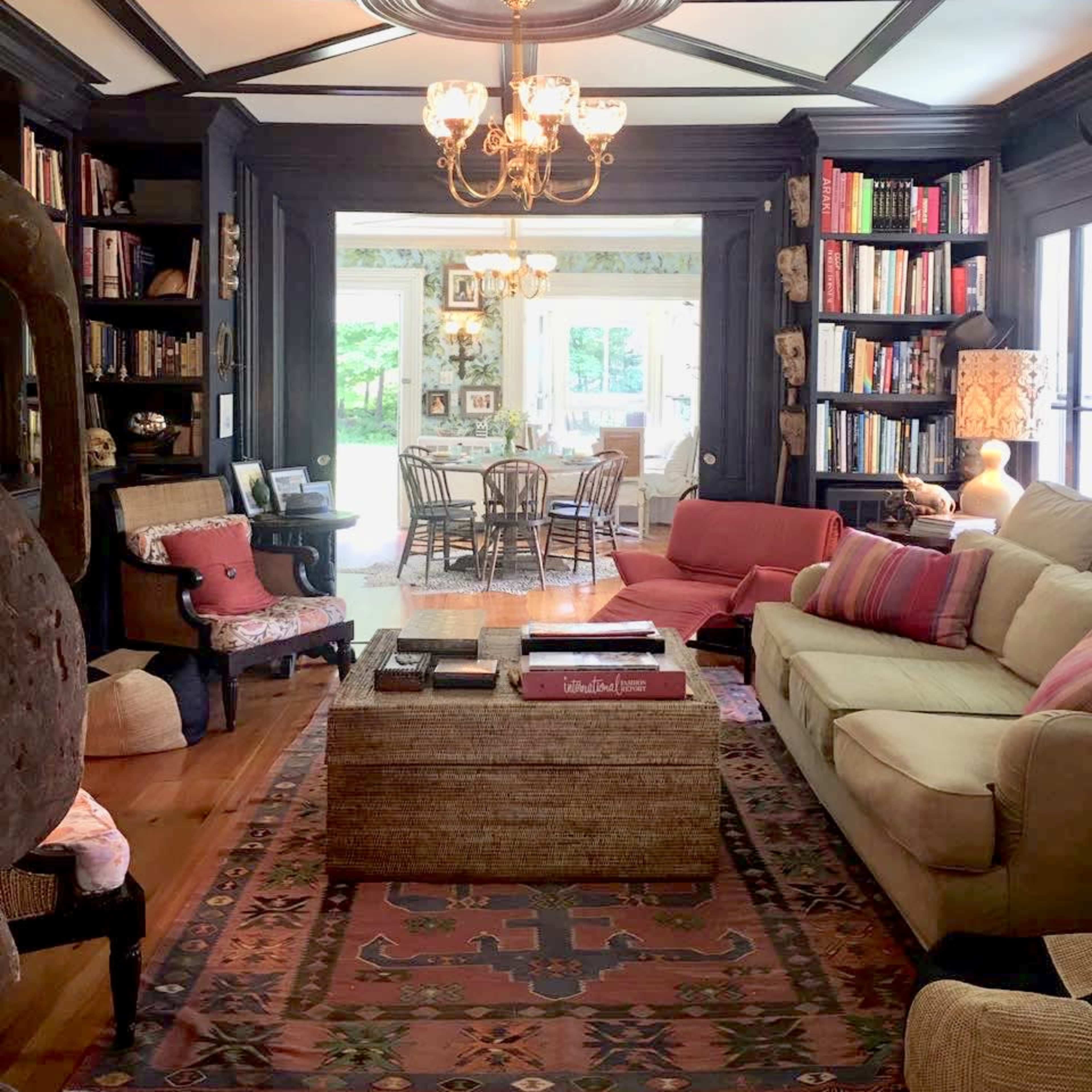 A cozy living room with dark wood accents, a chandelier, a large sofa, and bookshelves filled with books, leading to a bright dining area with a table and chairs visible through large windows.