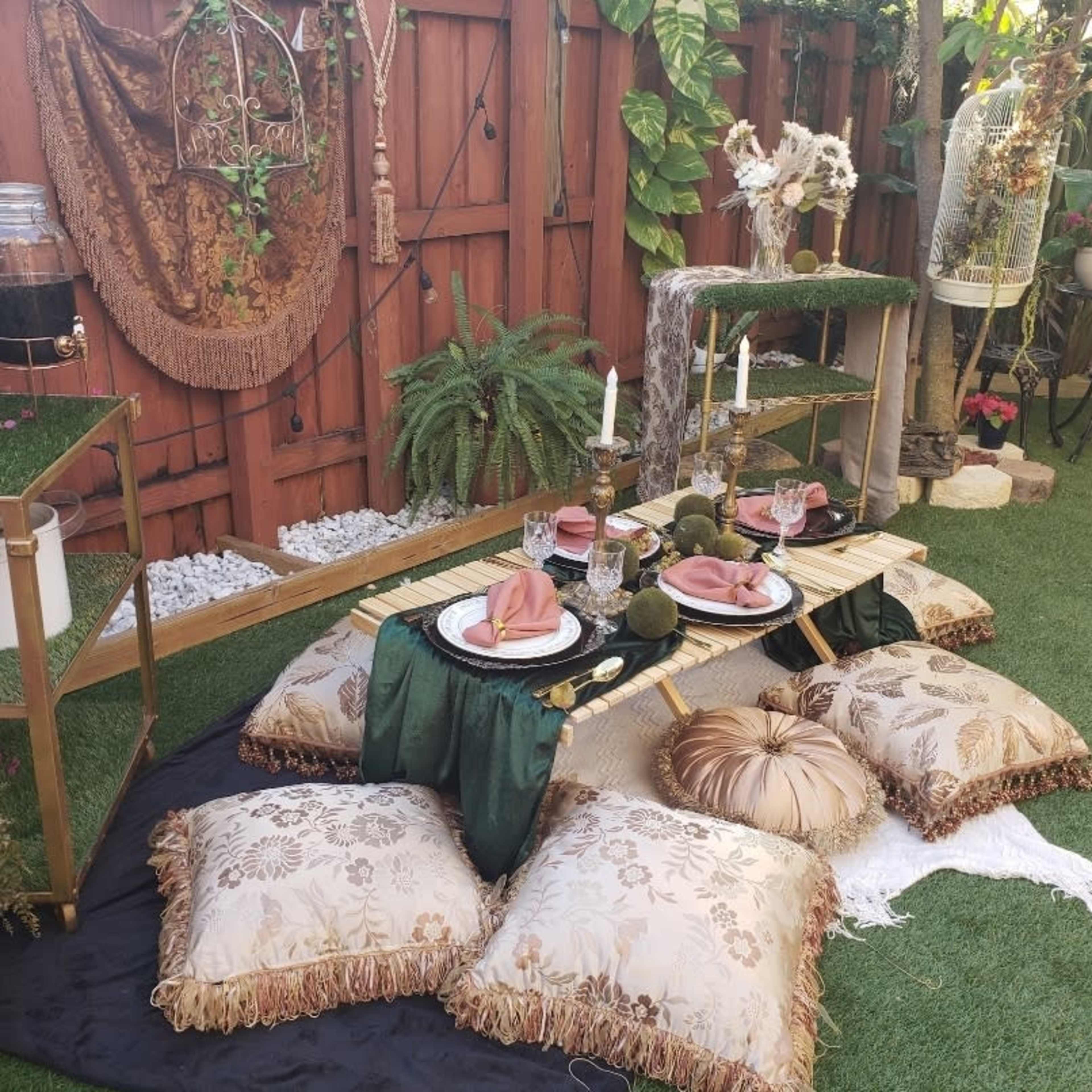 A low dining setup features a bamboo table adorned with dishes and cutlery, surrounded by decorative pillows on a grassy area against a wooden fence.