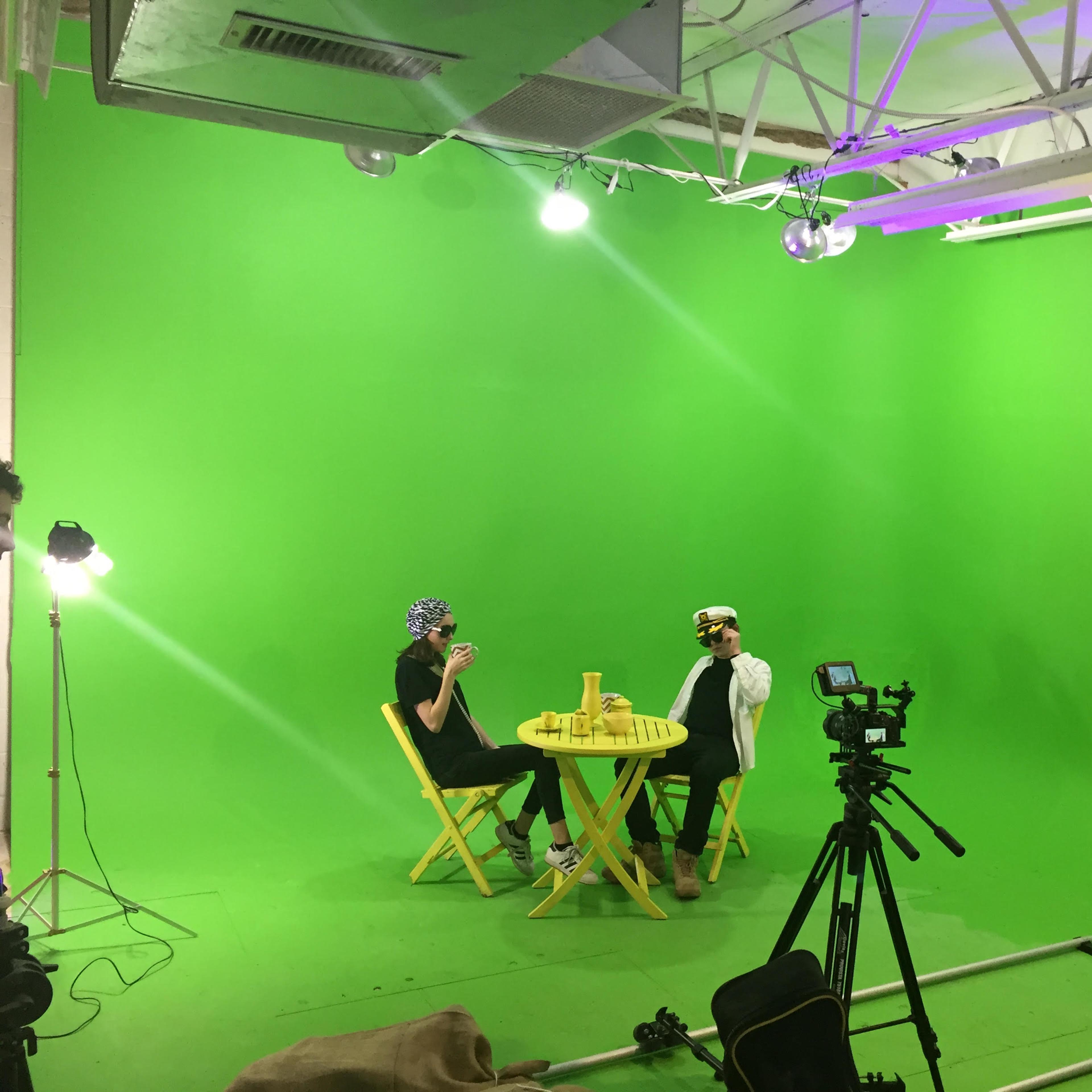 Two people are seated at a table in a green-screen studio, surrounded by cameras and lighting equipment.