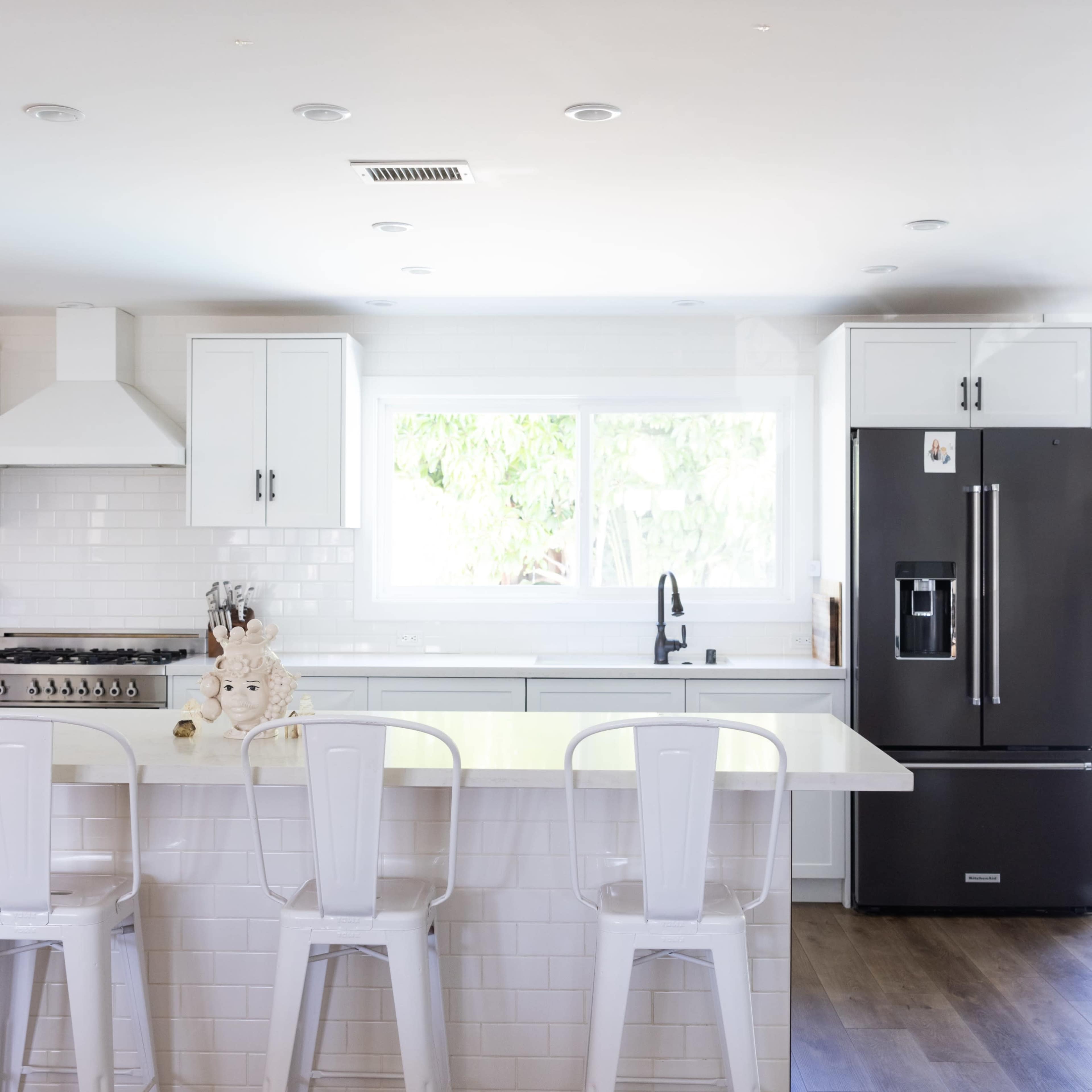 A modern kitchen features white cabinetry, a large island with three metal stools, and a black refrigerator, all illuminated by natural light from a window.