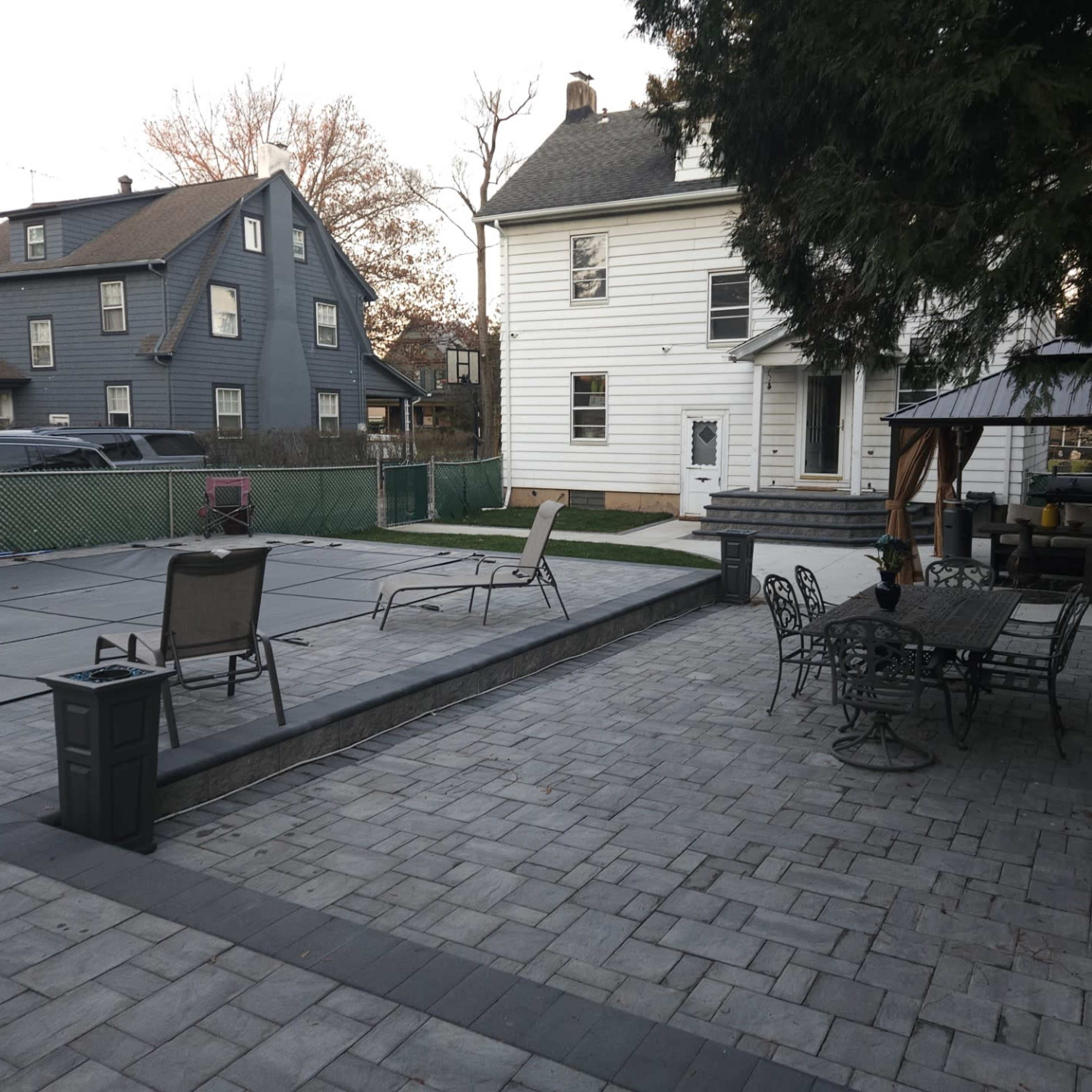 A backyard patio area with a pool covered and surrounded by paver stones, featuring lounge chairs, a dining set, and a gazebo.