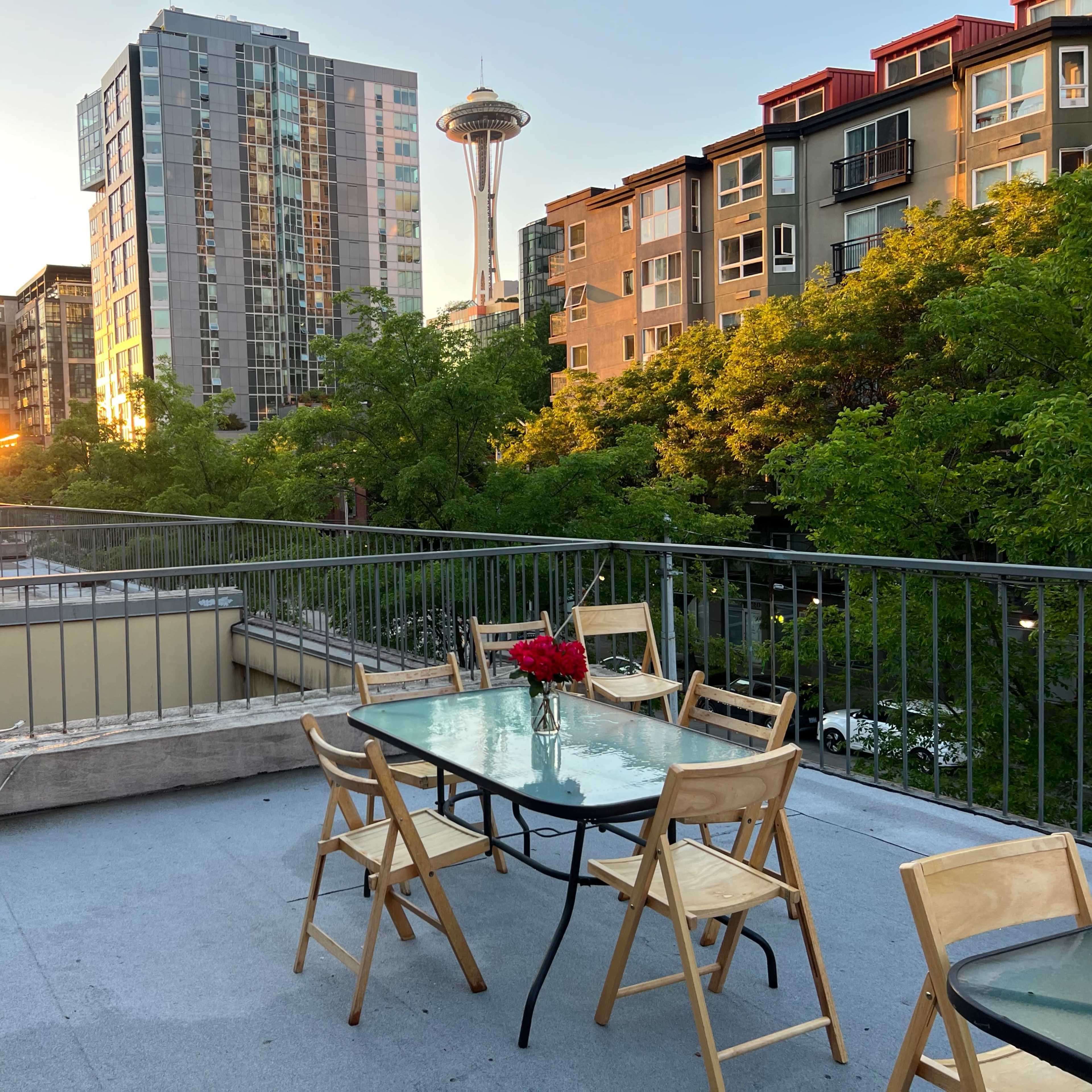 A rooftop patio with a glass table and wooden chairs overlooks a cityscape at sunset, featuring the Space Needle in the background.