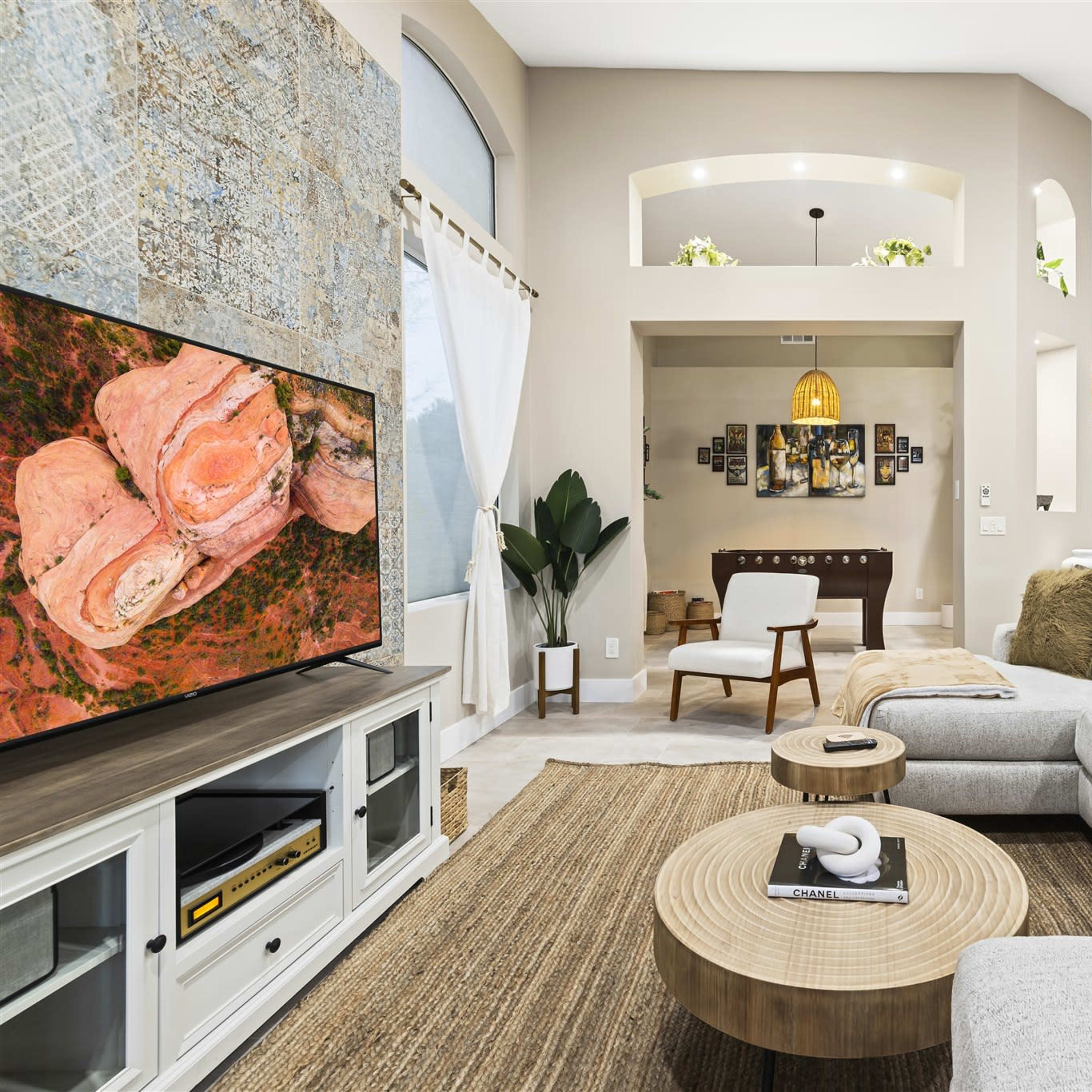 A modern living room features a large sectional sofa, a circular coffee table, a TV displaying artwork, and an open layout leading into a dining area.