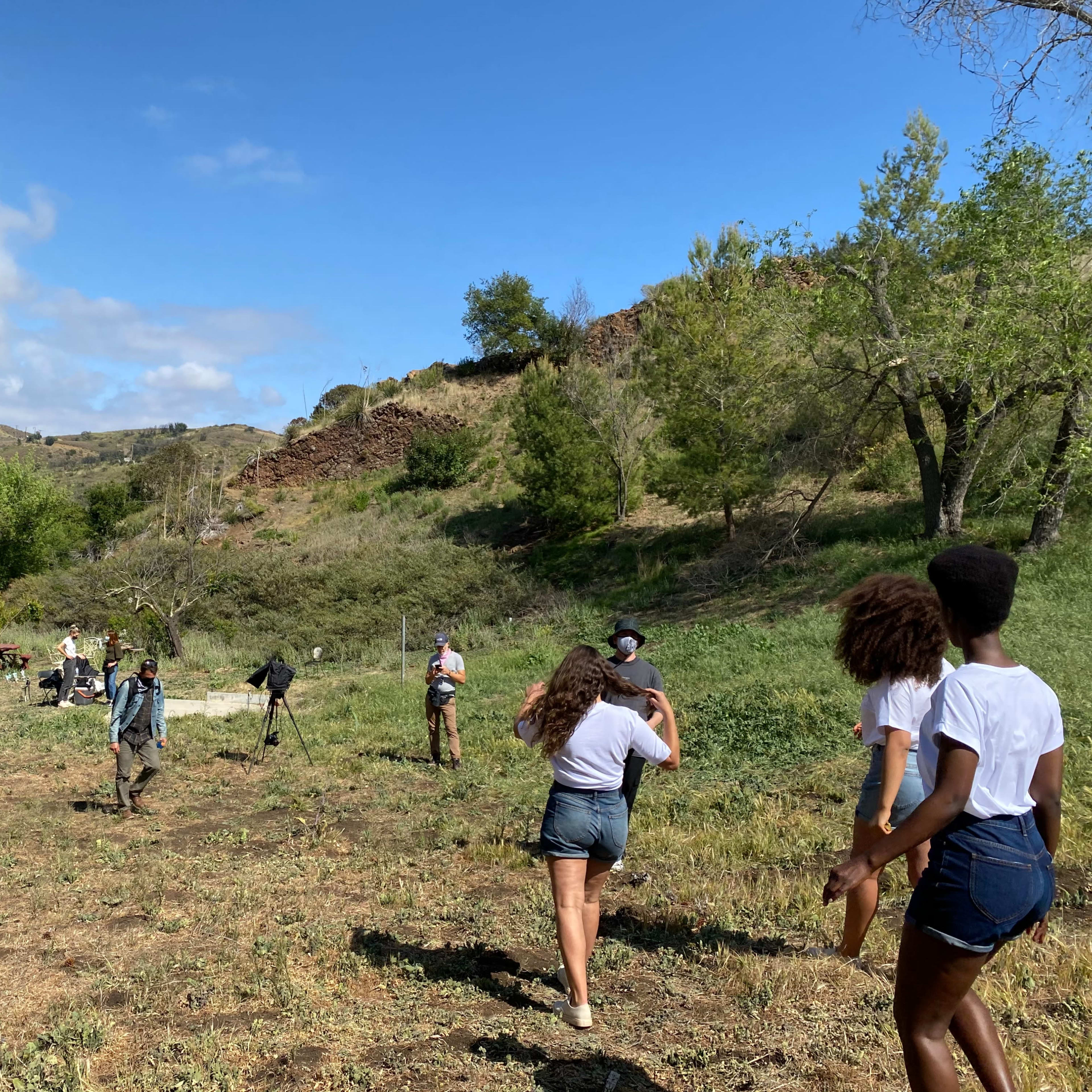 A group of people is walking across a grassy field with trees and hills in the background.