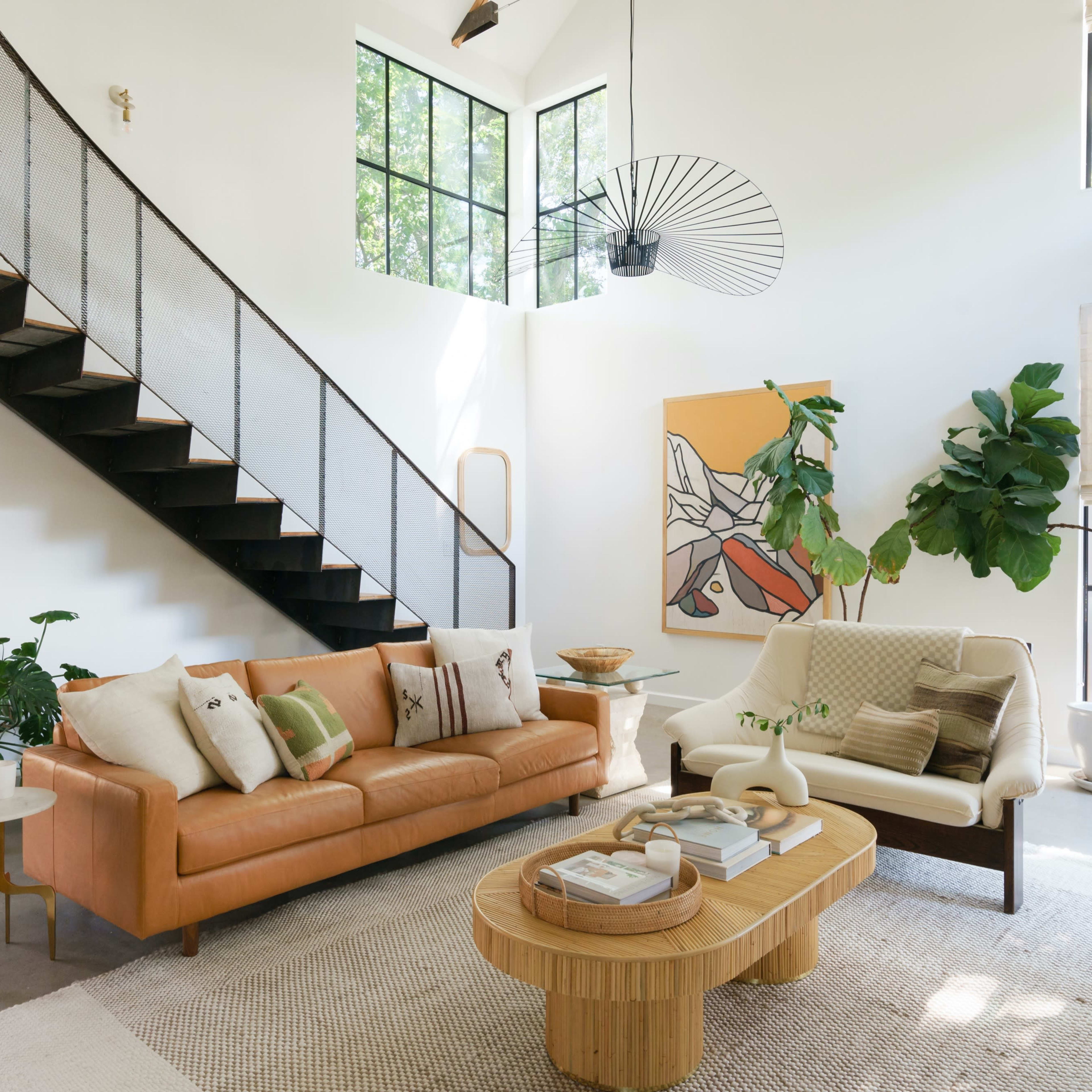 A living room features a staircase, two sofas, a round coffee table, and indoor plants, with large windows allowing natural light to enter.