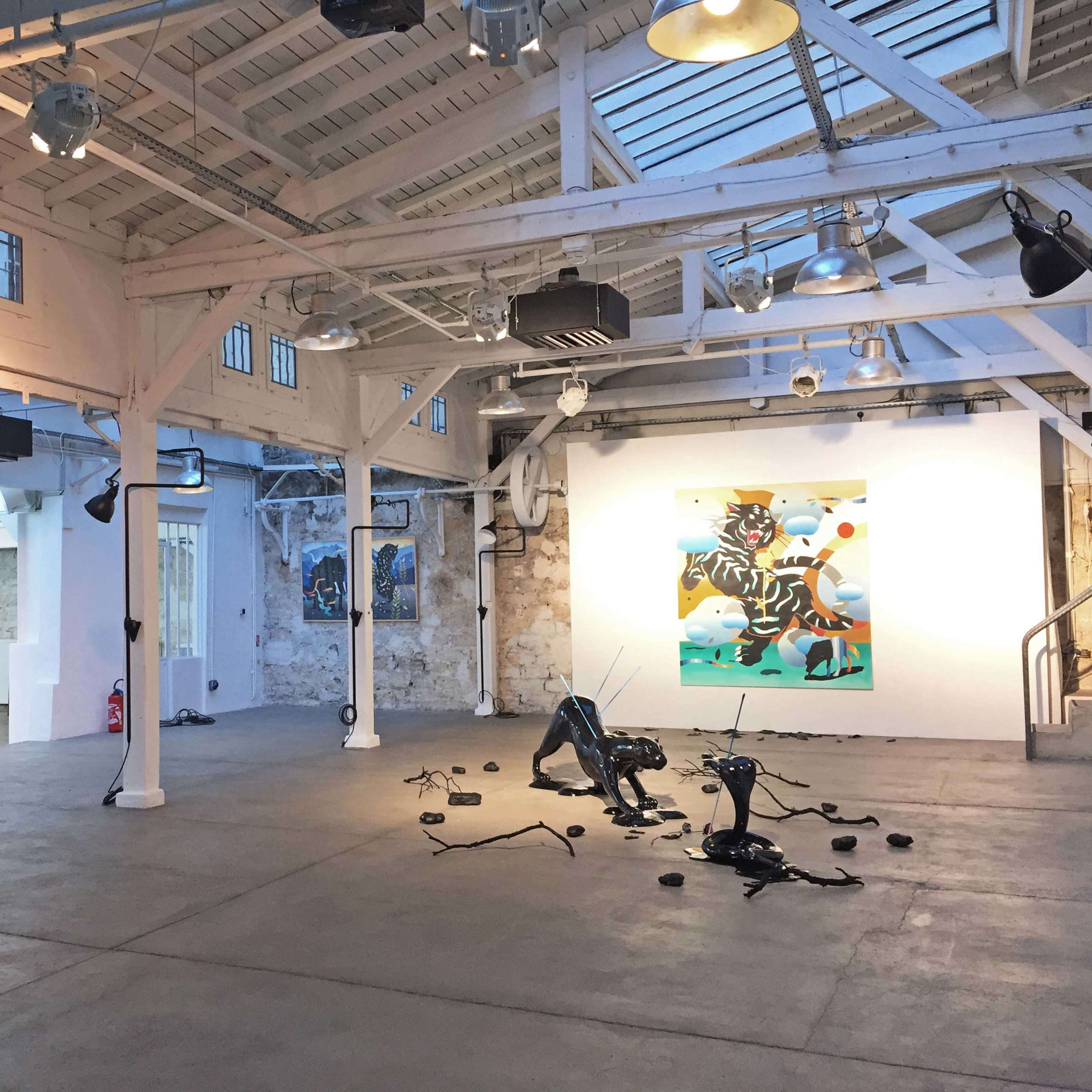 A bright art gallery featuring white walls, exposed beams, and scattered contemporary art pieces, including a large painting and various sculptural installations on the floor.